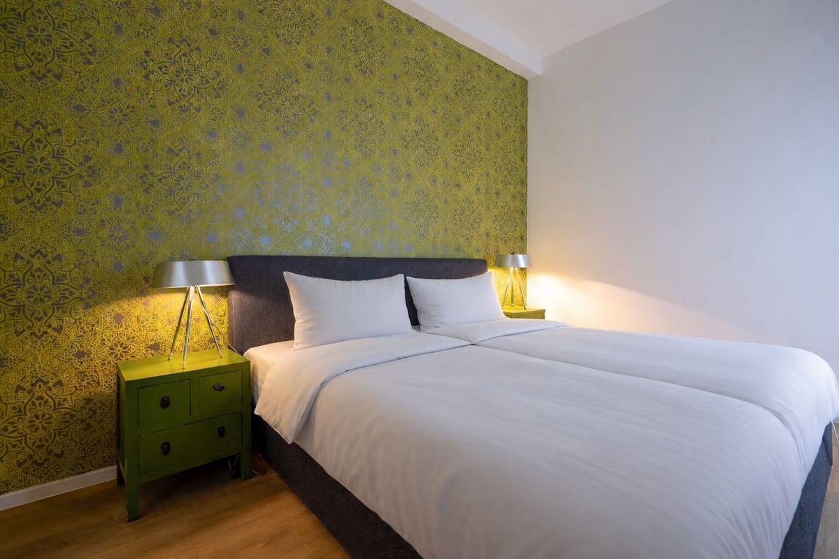 Een net opgemaakt bed met witte lakens, een nachtkastje in het groen met een lamp, en behang met een patroon in een hotelkamer.