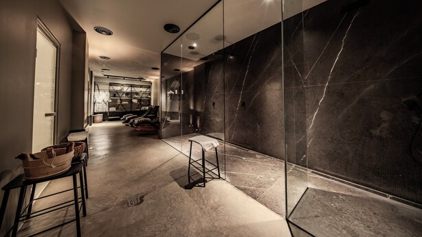 Een moderne hotelgang met een glazen scheidingswand, een zithoek met een bank en een uitzicht op een kamer met een bed en een grote spiegel.