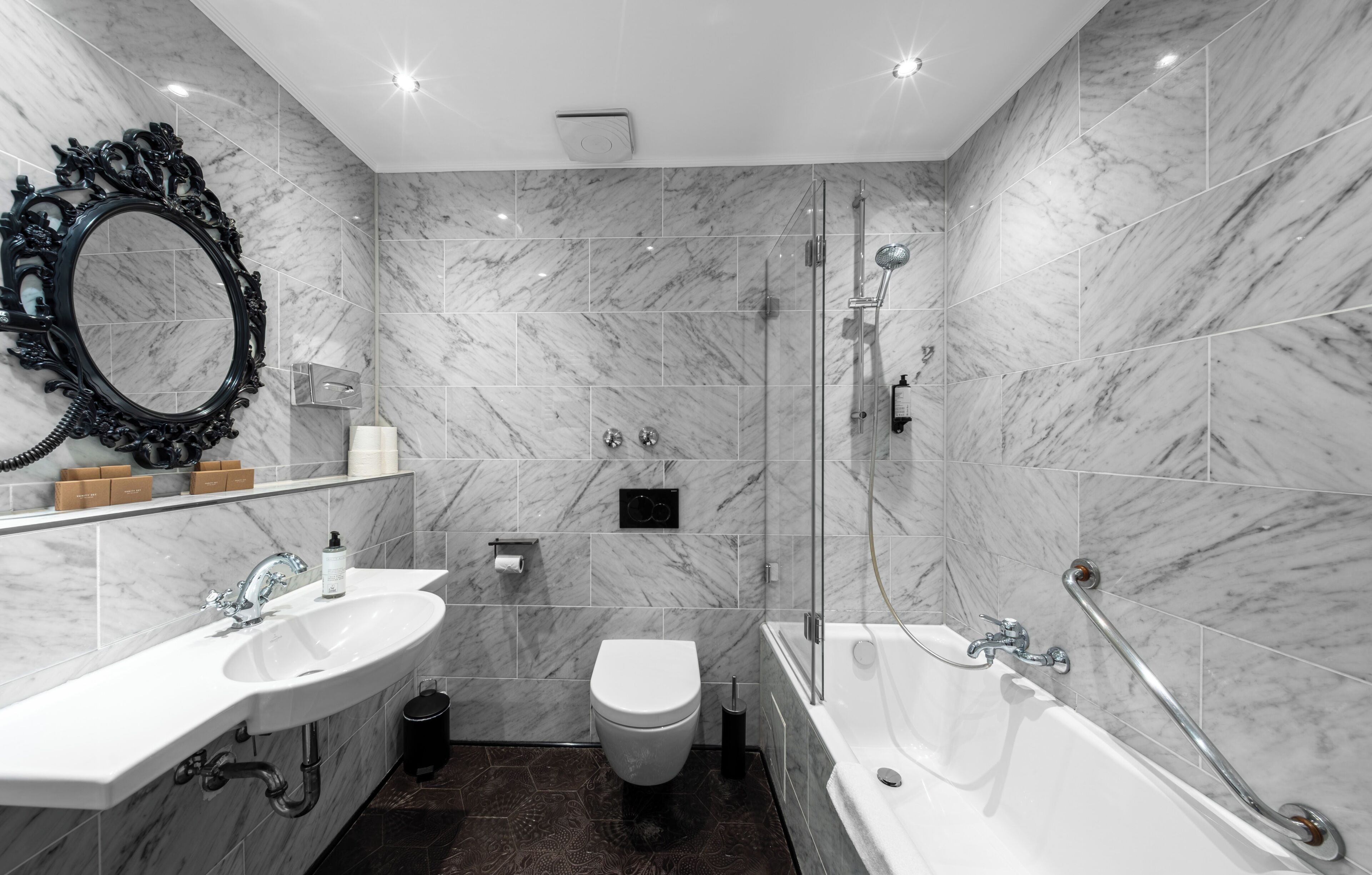 A modern bathroom with a marble sink, toilet, and bathtub, equipped with a handheld showerhead and grab bars.