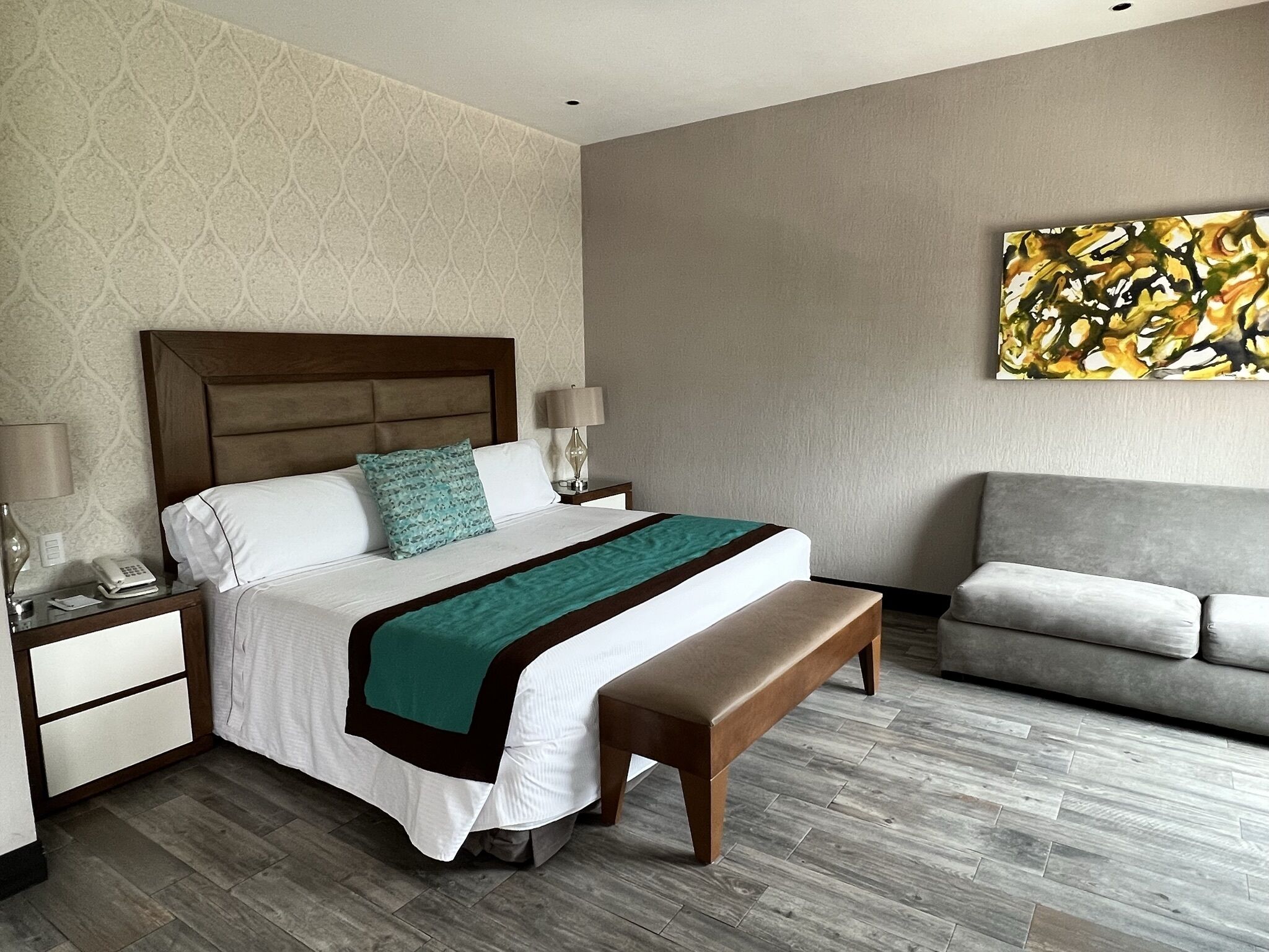 A hotel room with a bed, bedside tables, a bench, a sofa, and a painting on the wall.