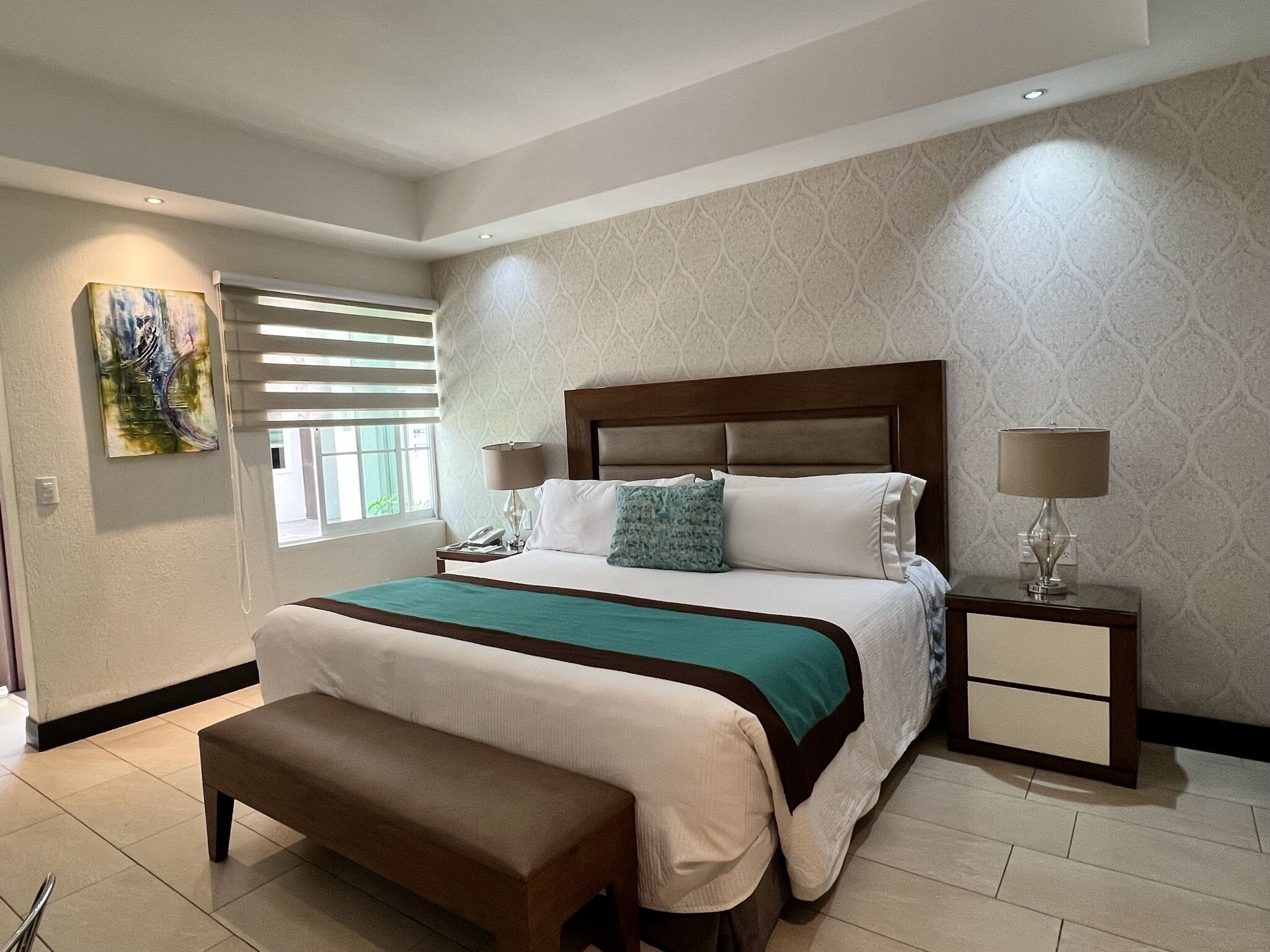 A bedroom with a bed, bedside tables, a bench, a painting, and a window with blinds.