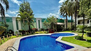A swimming pool with a 140 m² area, surrounded by a landscaped garden with palm trees and a building with green and white walls in the background. - Áurea Hotel & Suites (Zapopan)