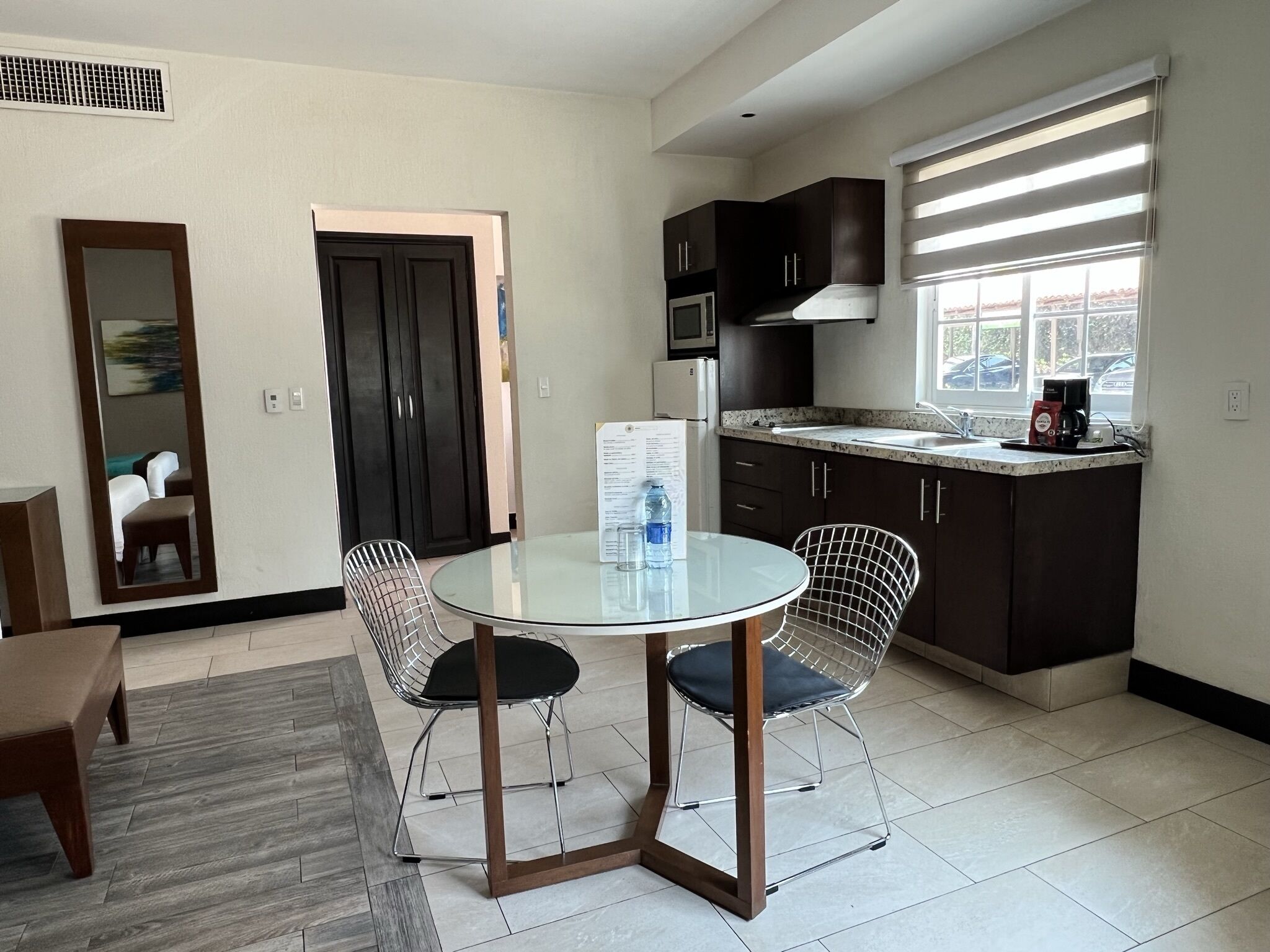 A modern kitchen with dark cabinets, a microwave, and a coffee maker. There is a round glass table with two wireframe chairs.