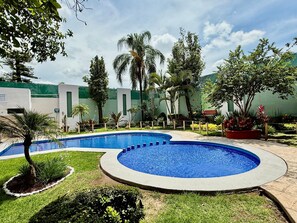 A swimming pool with a curved design, surrounded by well-maintained grass and various plants. - Áurea Hotel & Suites (Zapopan)