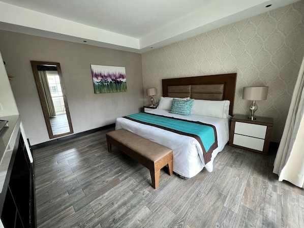 A modern hotel room with a large bed, a wooden bench, a nightstand with a lamp, and a painting on the wall. - Áurea Hotel & Suites (Zapopan)