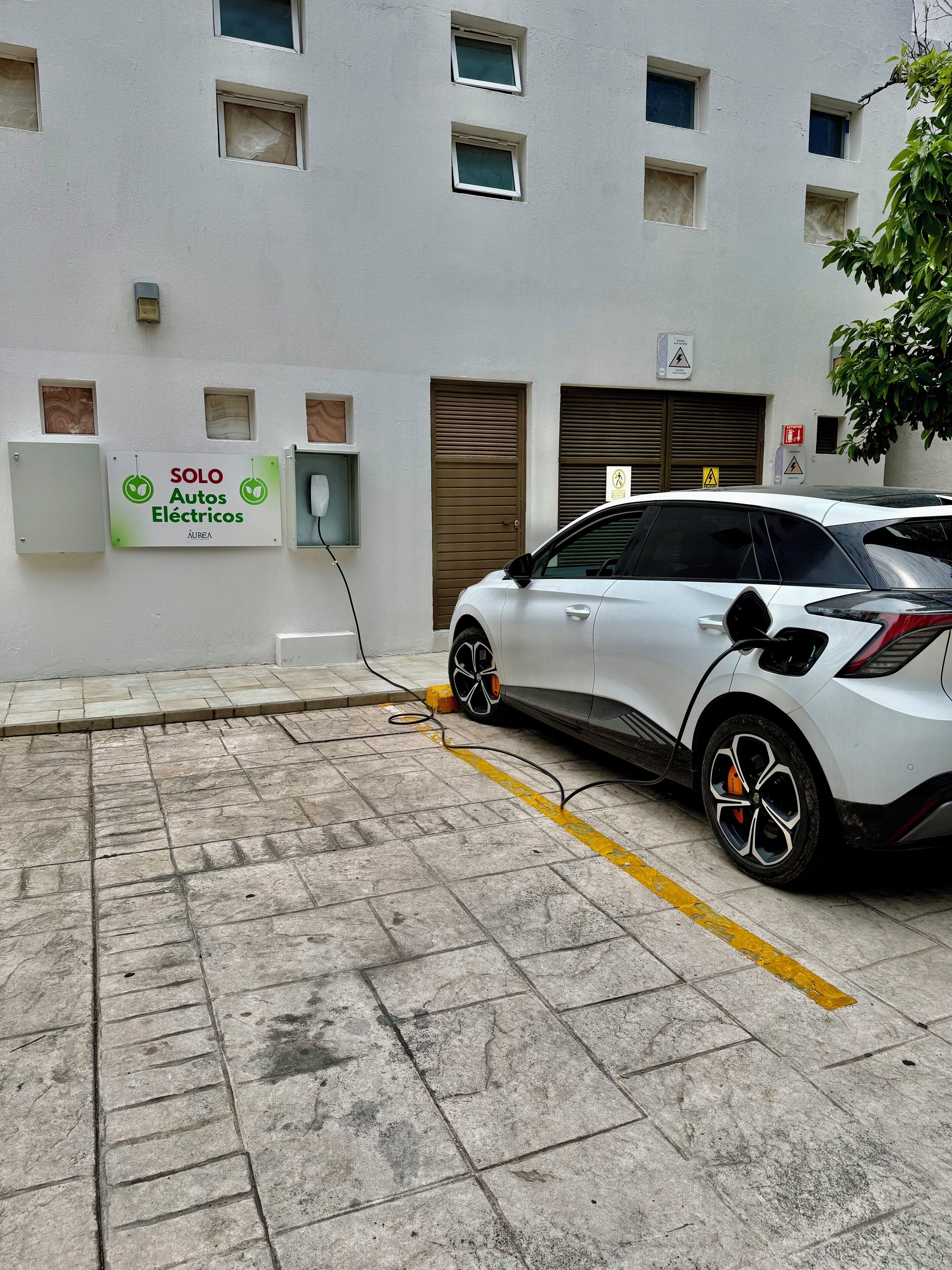 A white electric car charging at a station with a sign reading "SOLO Autos Eléctricos" on the wall.