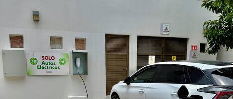 A white electric car charging at a station with a sign reading "SOLO Autos Eléctricos" on the wall.