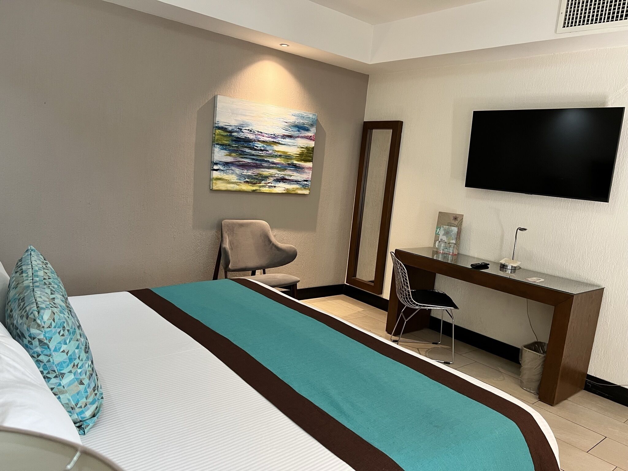 A hotel room with a bed, a desk, a chair, a TV, and a painting on the wall.