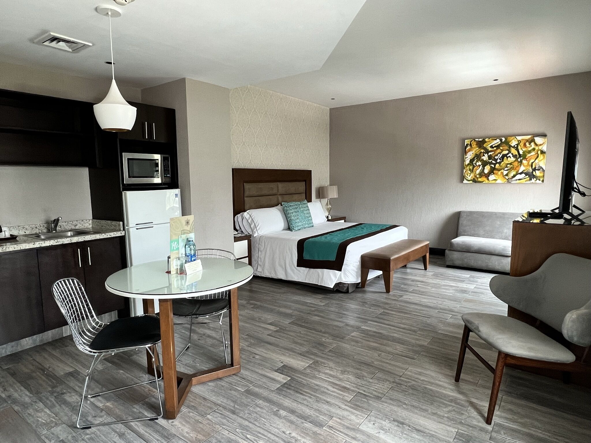 Una habitación de hotel moderna con kitchenette, una cama con cabecera, una mesita de noche, una silla, un sofá y un cuadro en la pared.