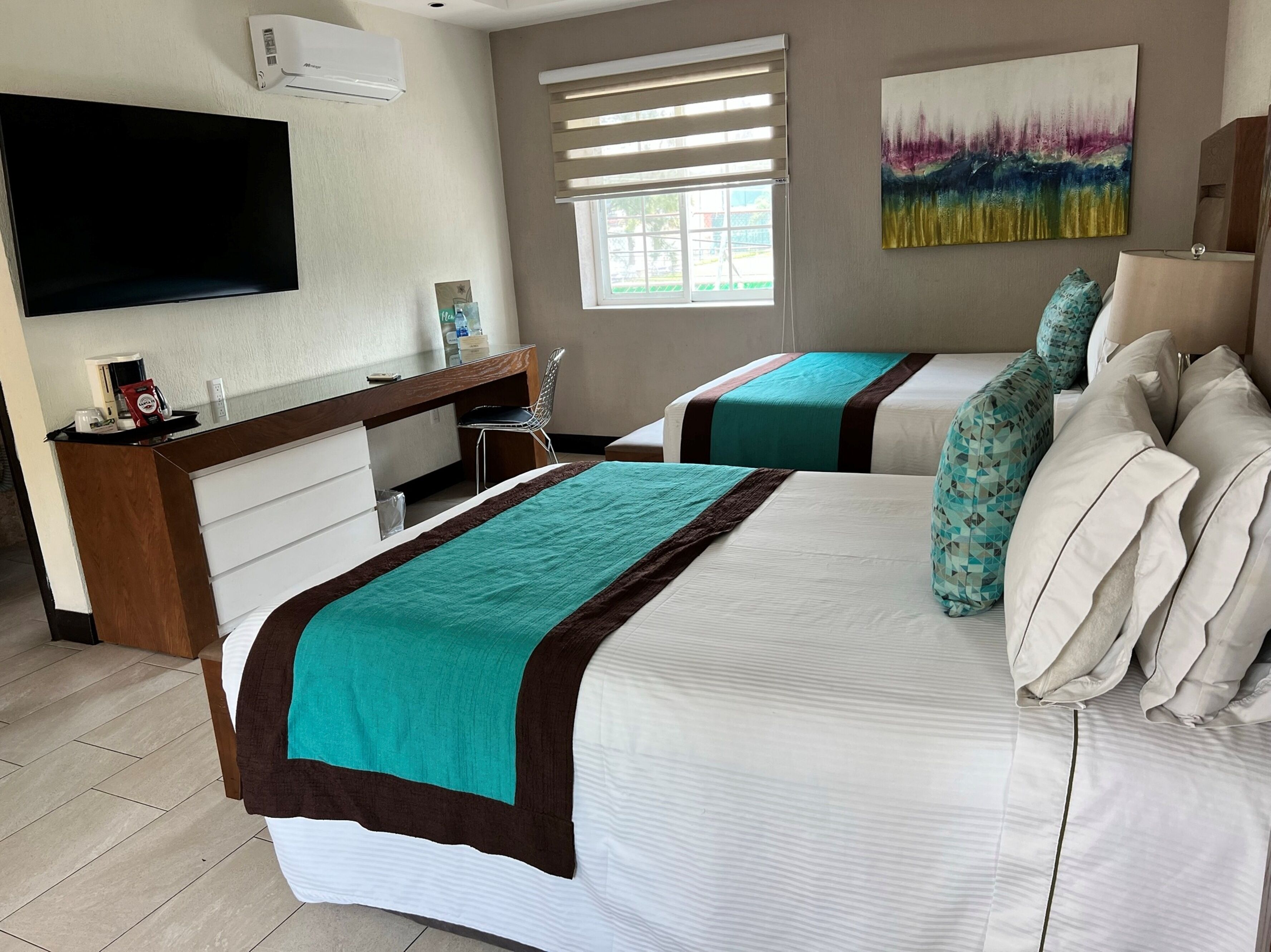 A hotel room with two beds, a desk with a chair, a flat-screen TV, and a window with blinds.
