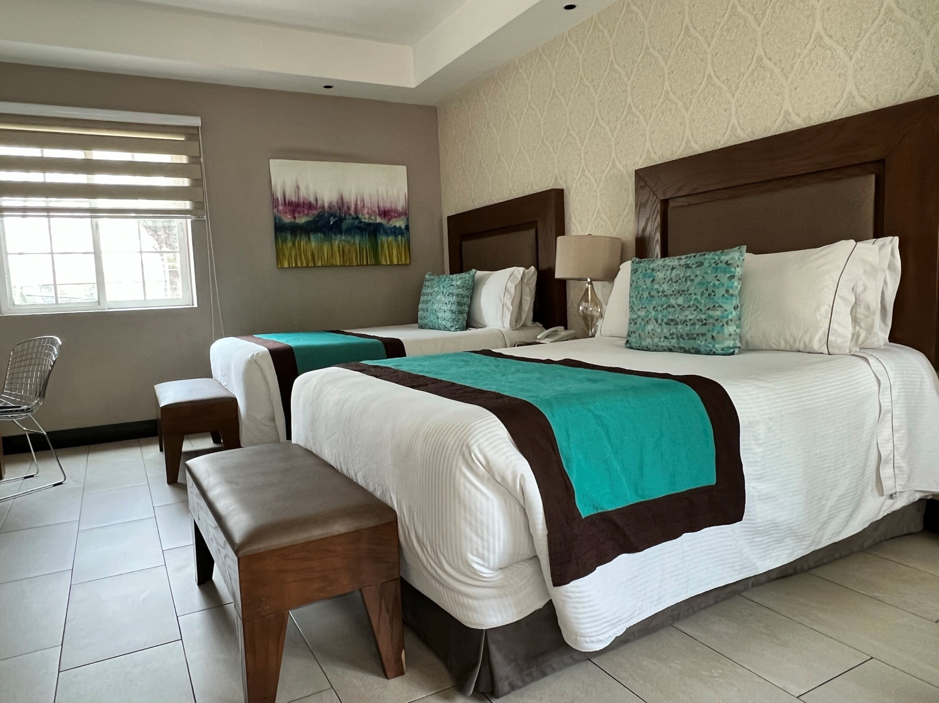A hotel room with two beds, a bench, a chair, a painting, and a window with blinds.