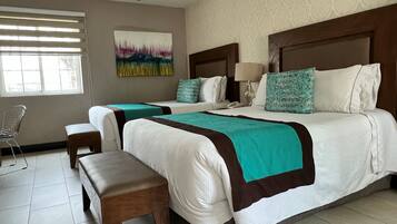 A hotel room with two beds, a bench, a chair, a painting, and a window with blinds.