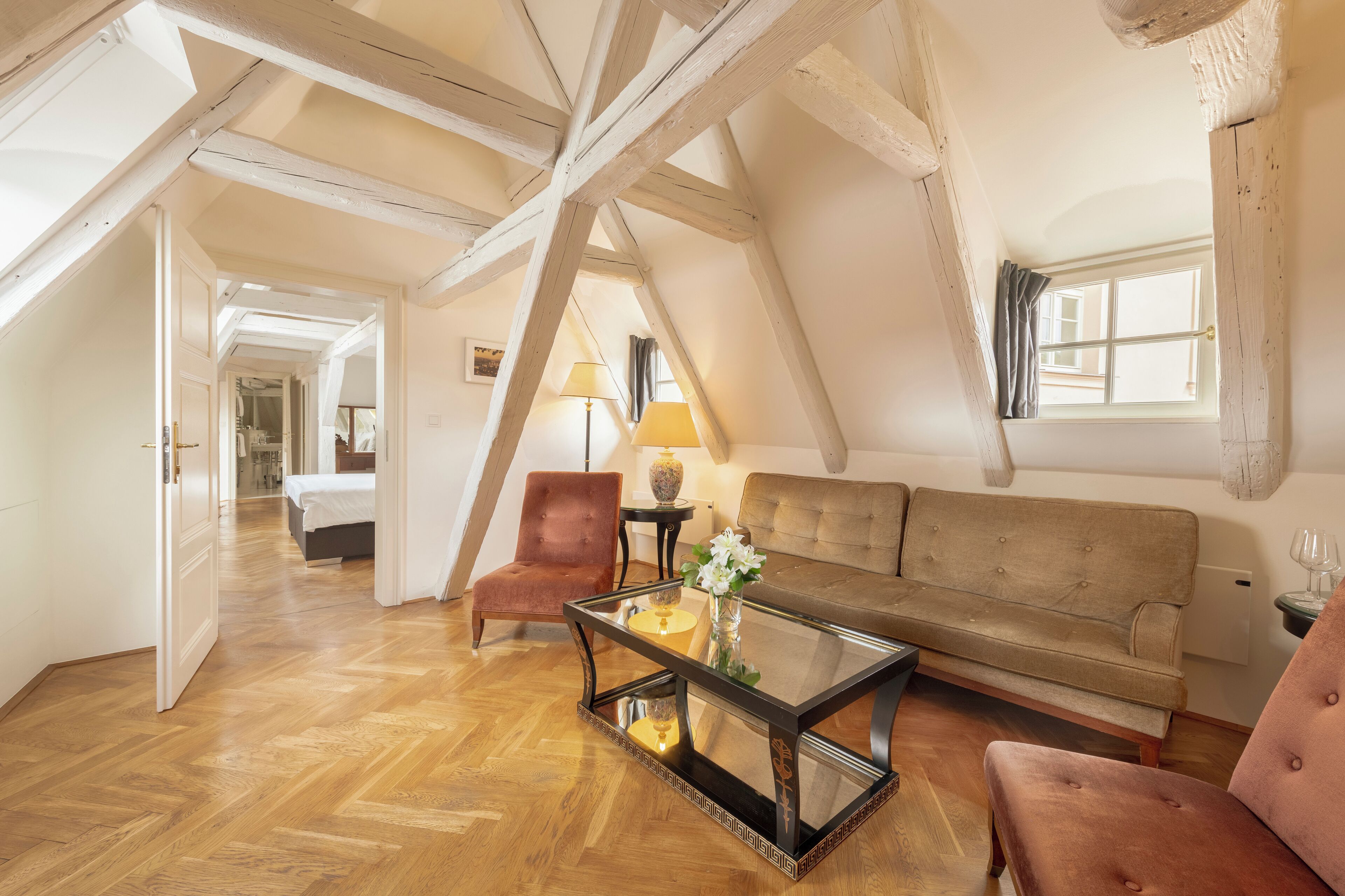 Um quarto estilo loft com vigas de madeira aparentes, uma mesa de centro de vidro, um sofá e uma poltrona.