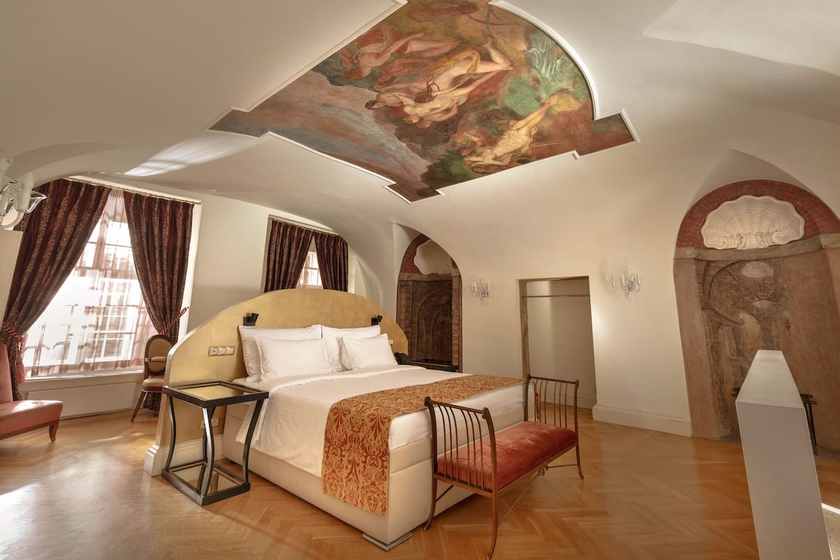 A spacious bedroom with a large bed, a desk, and a chair. The room has a high ceiling with a mural, and there are windows with curtains.