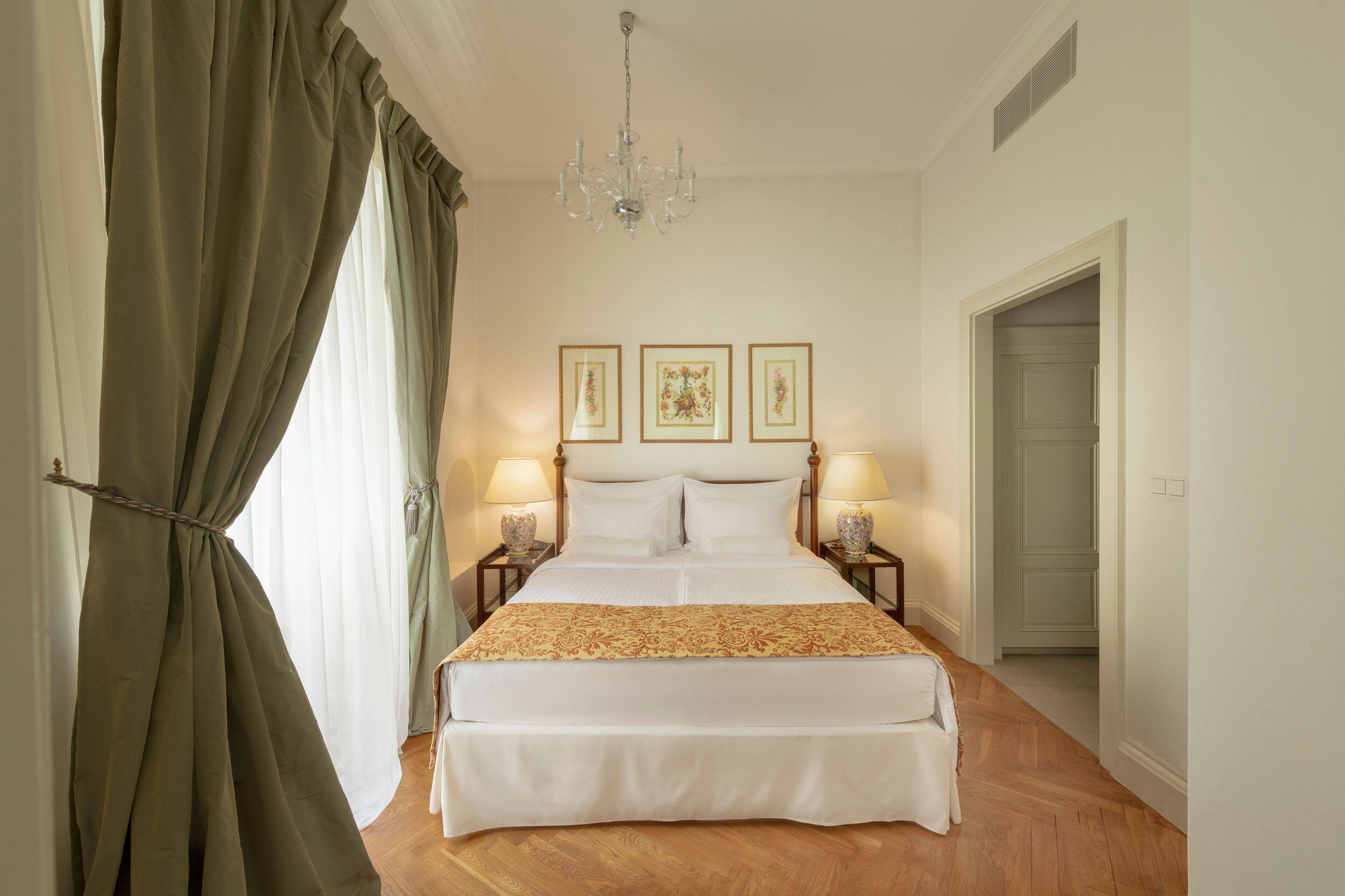 Una cama bien hecha con edredón blanco y una manta decorativa, flanqueada por dos mesitas de noche con lámparas, en una habitación con suelo de madera y un candelabro.