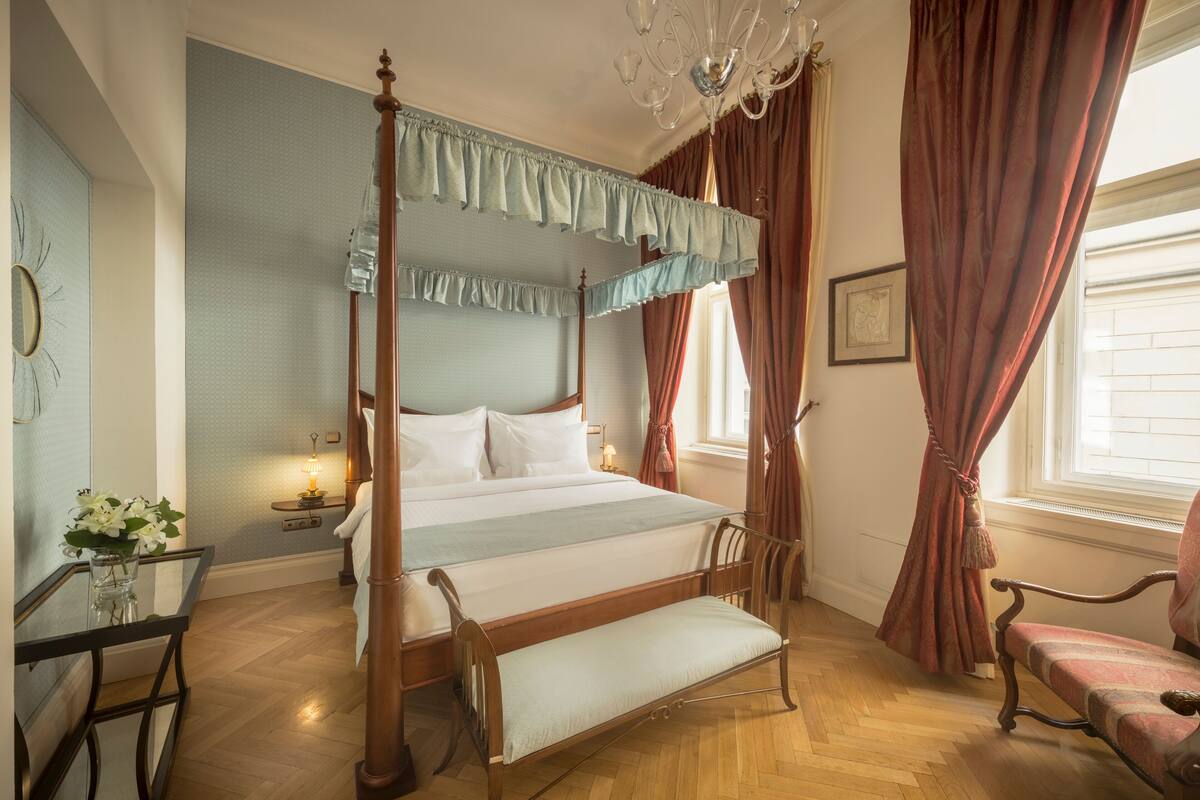 A four-poster bed with a wooden frame and white bedding, a chandelier, and a window with curtains.