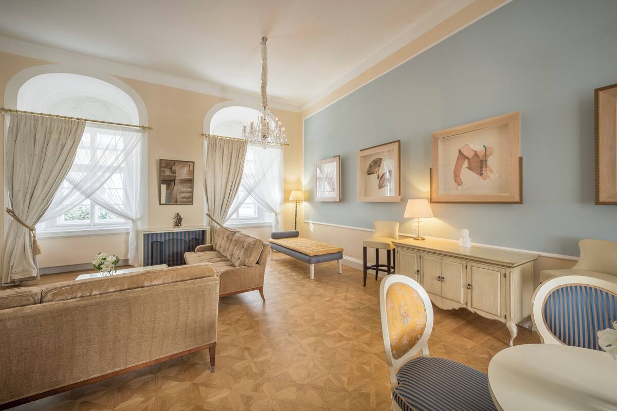 A room with a sofa, armchair, and a small table with a lamp. There is a window with curtains and a chandelier.