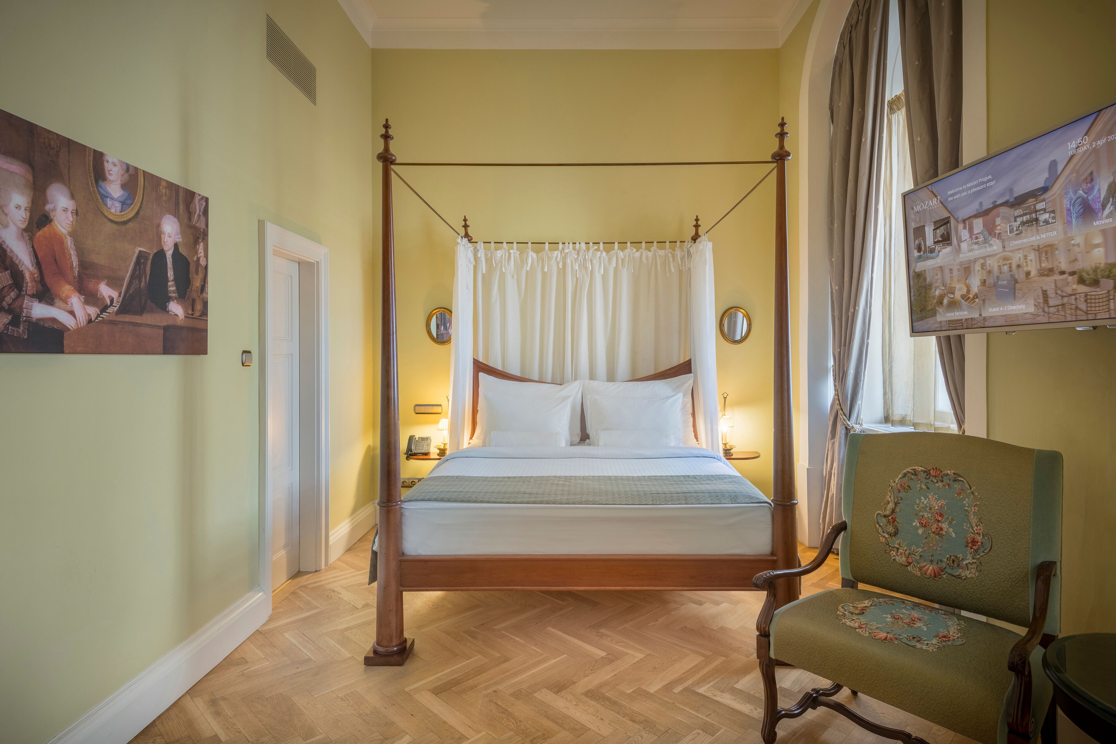 Una cama con dosel, suelo de madera, una butaca tapizada de verde y un cuadro en la pared.