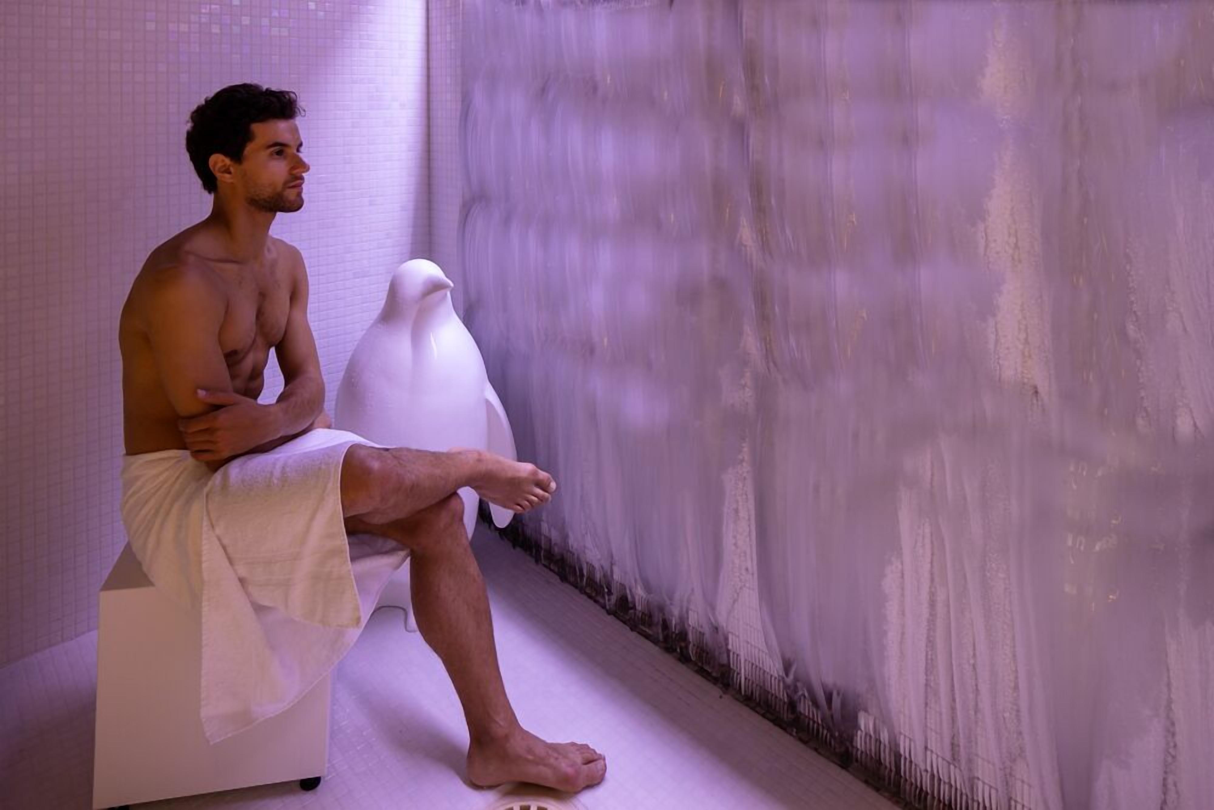 A person sitting on a white bench in a room with a textured wall and a floor drain.