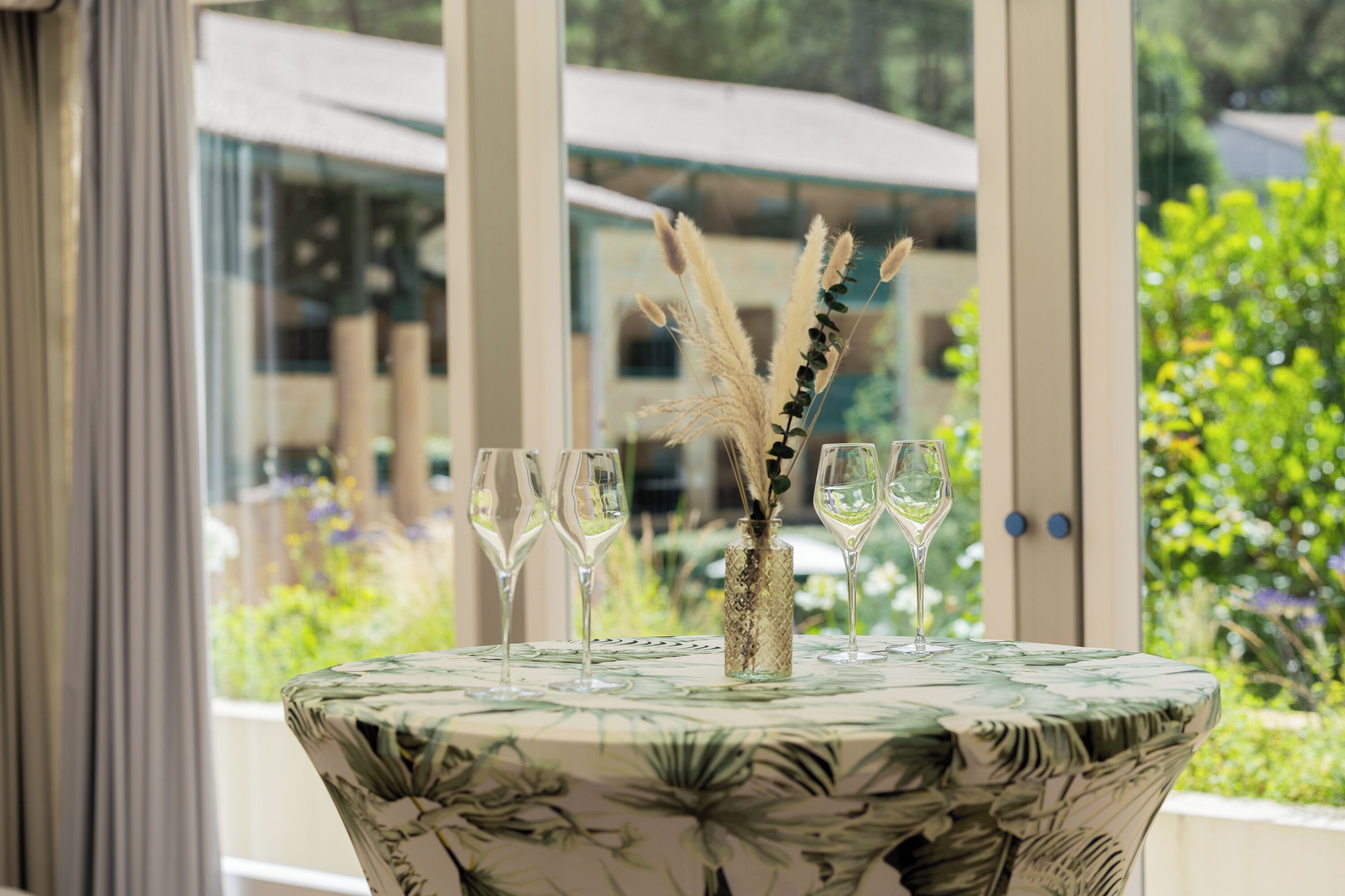 Uma mesa com toalha florida, um vaso com plantas secas e três copos de vinho.