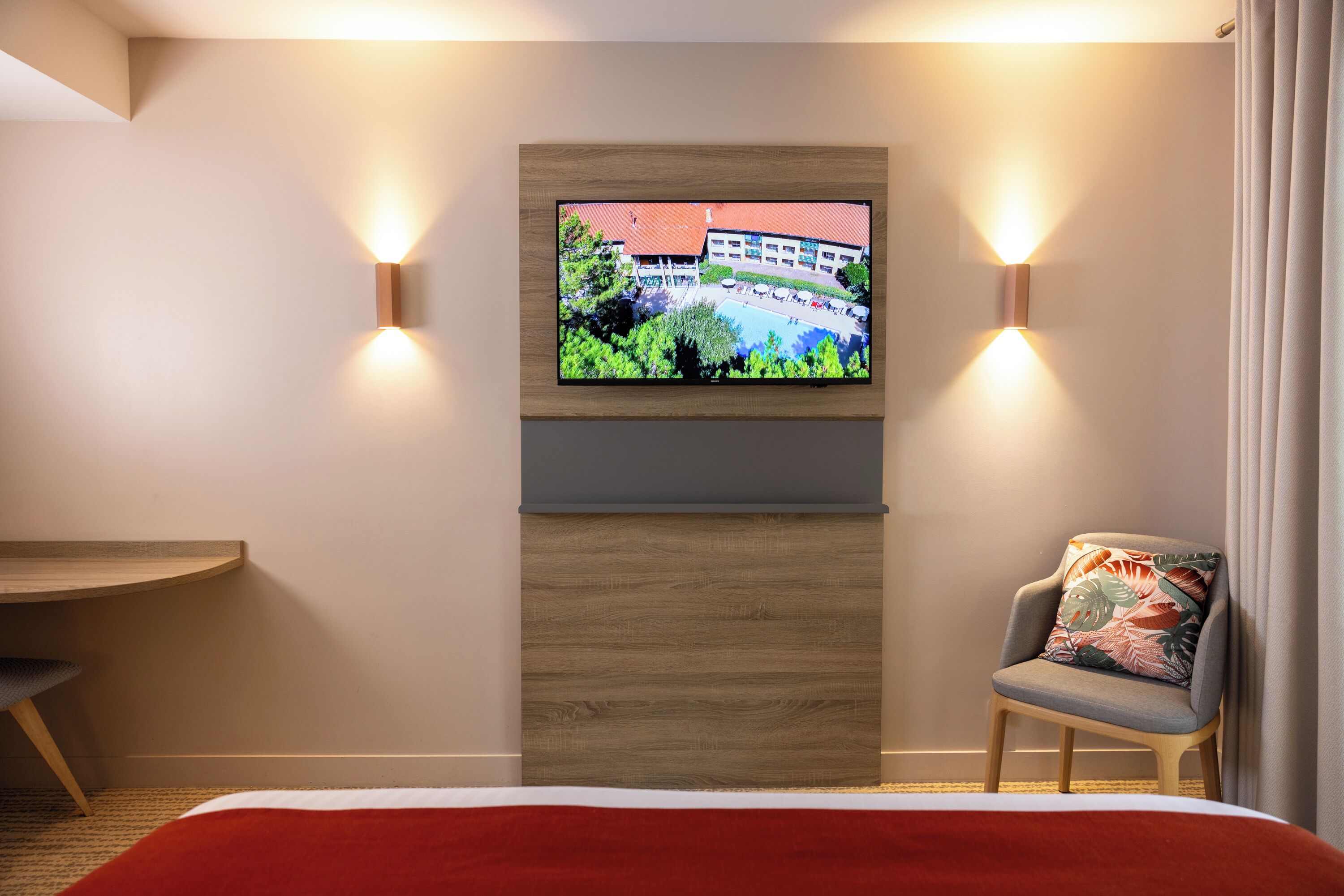 A hotel room with a bed, a TV mounted on the wall, a chair, and a wooden headboard.