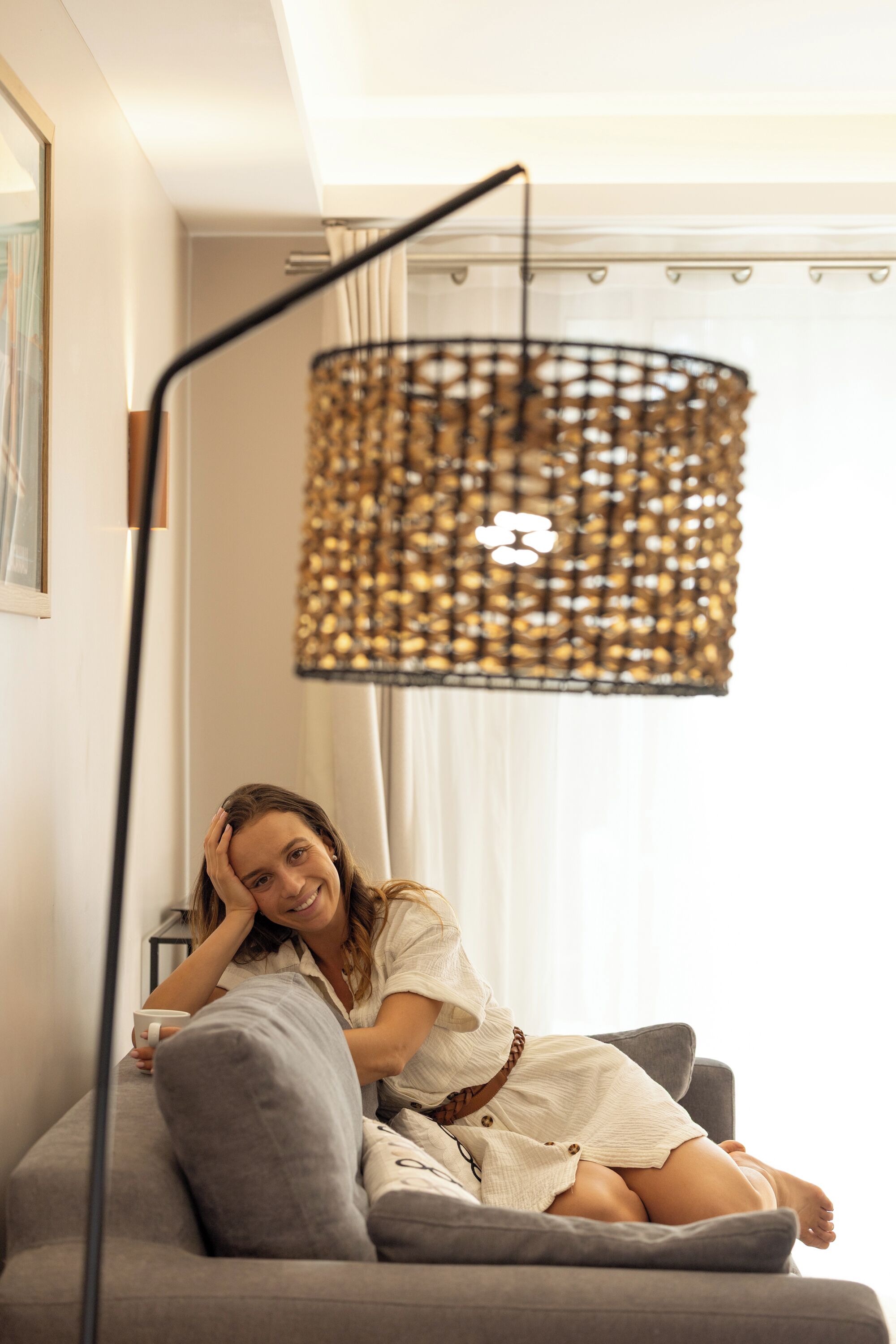 Une personne est assise sur un canapé gris dans une pièce où l’on aperçoit une lampe en osier, un tableau encadré au mur et une fenêtre avec des rideaux.