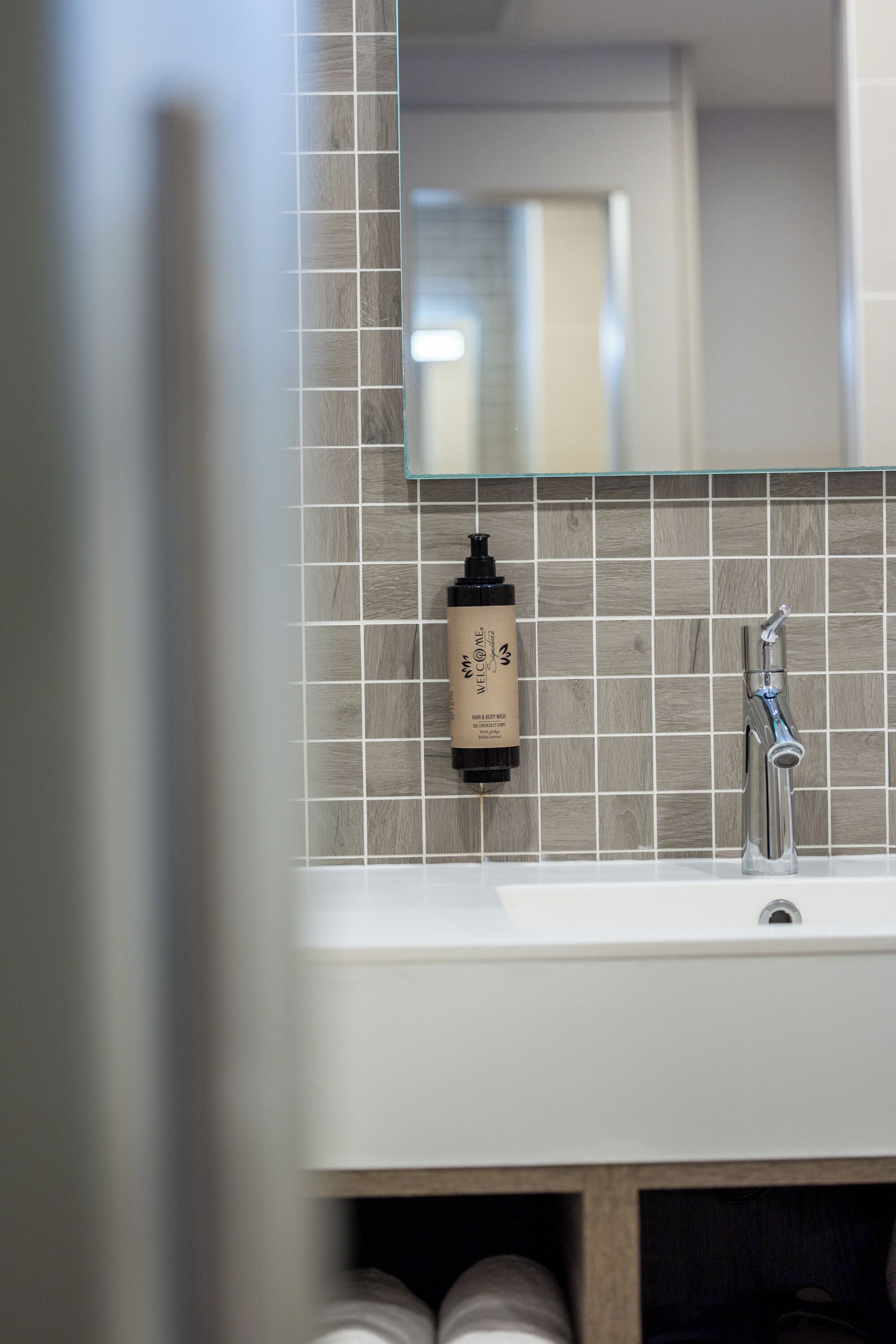 Une salle de bain avec un mur recouvert de carreaux derrière le lavabo, un lavabo, un miroir et une bouteille de shampoing.