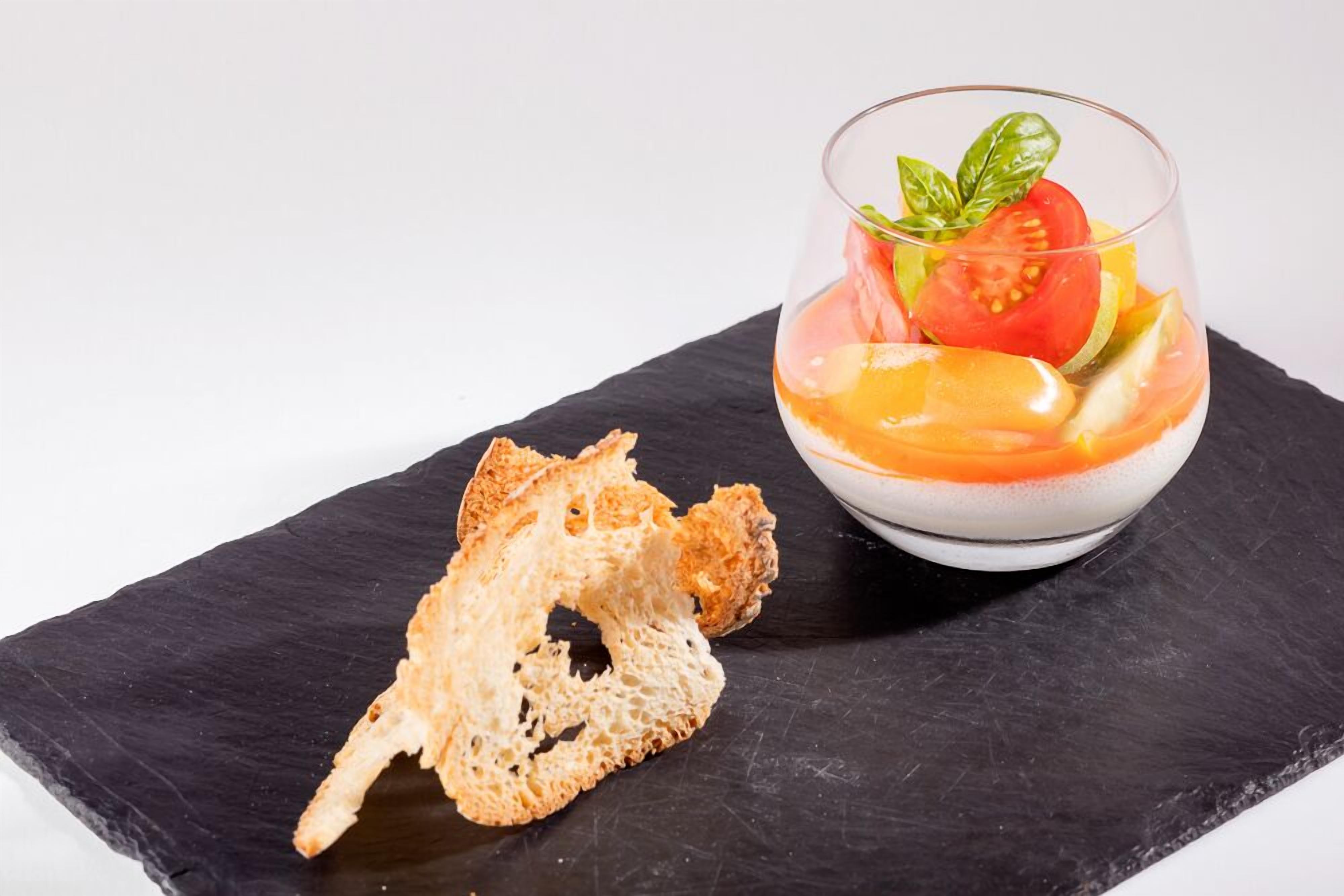 A dessert in a glass bowl with a basil leaf garnish, accompanied by a piece of bread on a slate board.