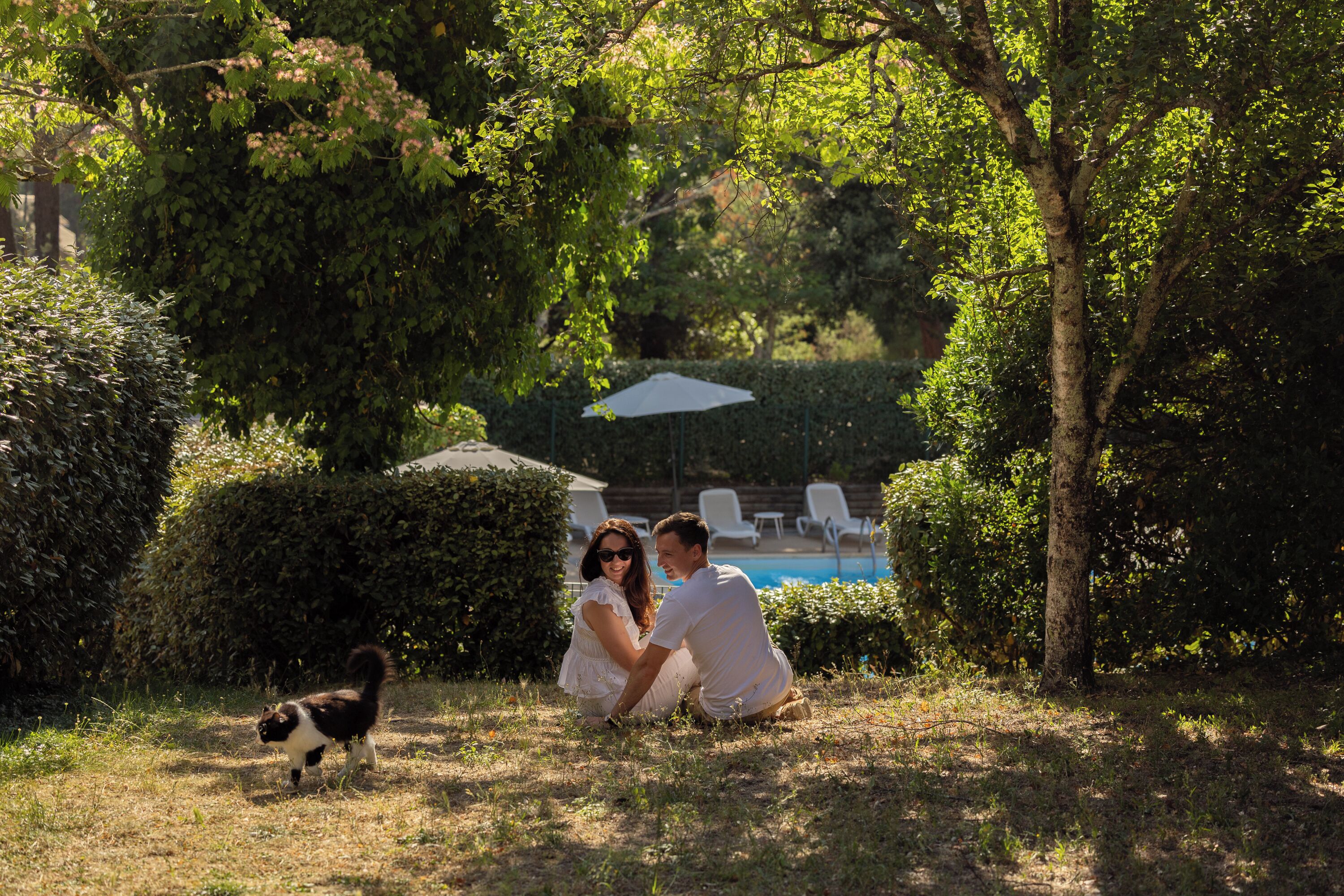 Un couple est assis sur l’herbe, près d’une piscine, entouré d’arbres et d’arbustes.