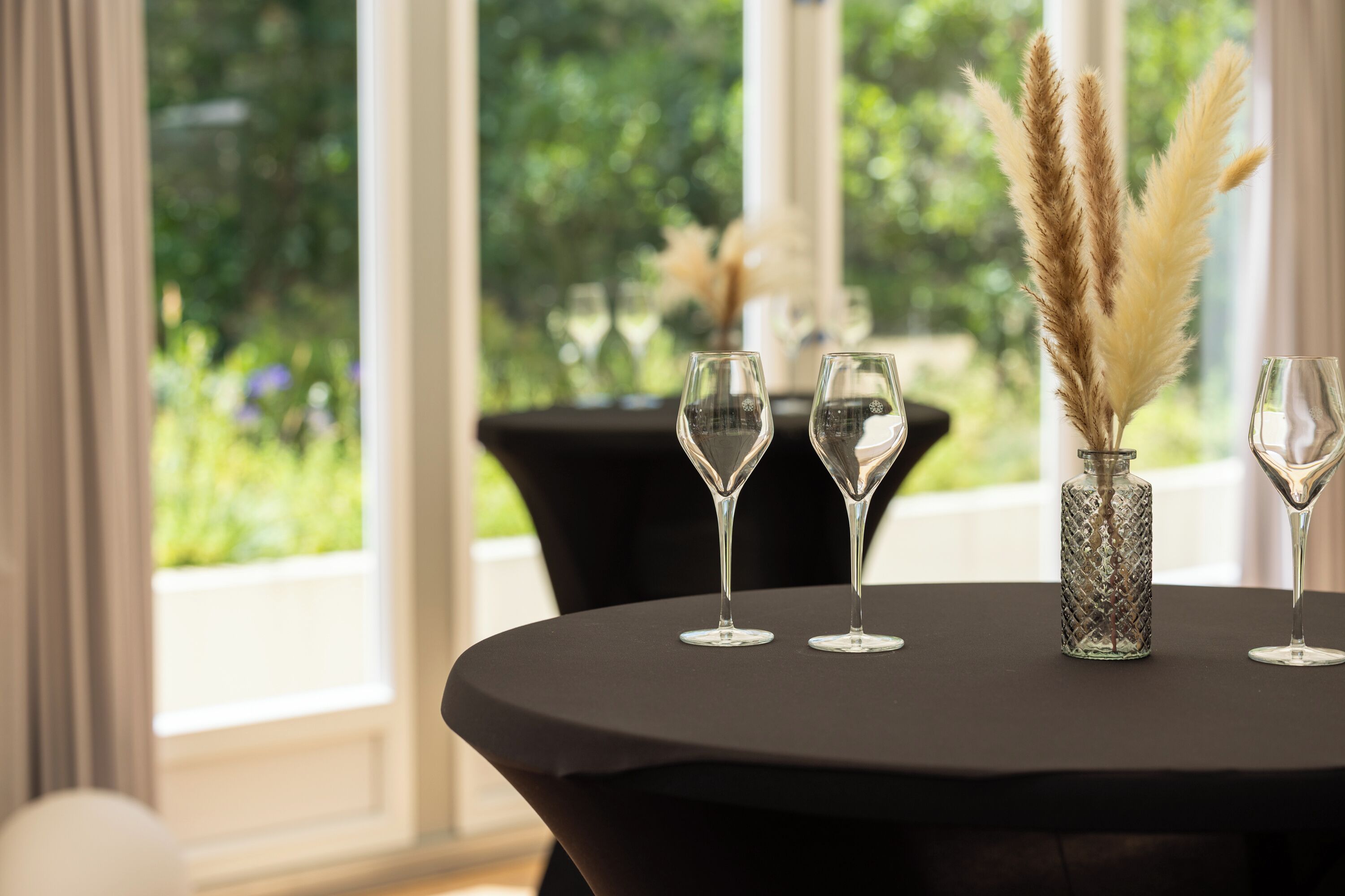 Une table avec trois verres à vin et un vase contenant des plumes d’herbe des pampas, le tout placé devant une fenêtre donnant sur un paysage verdoyant.