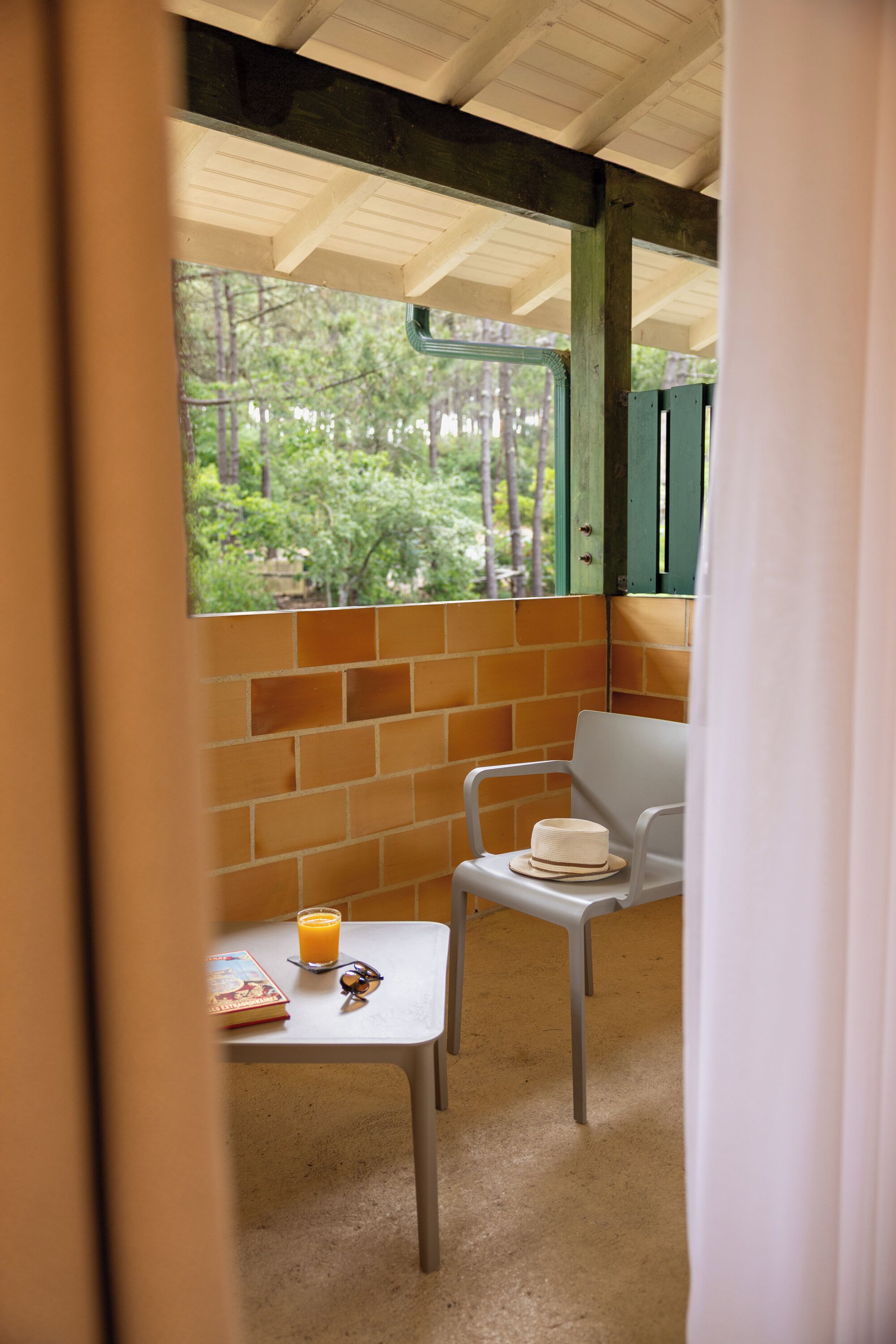 Una habitación pequeña con ventana, una mesa con un libro y un vaso de jugo de naranja, y una silla blanca.