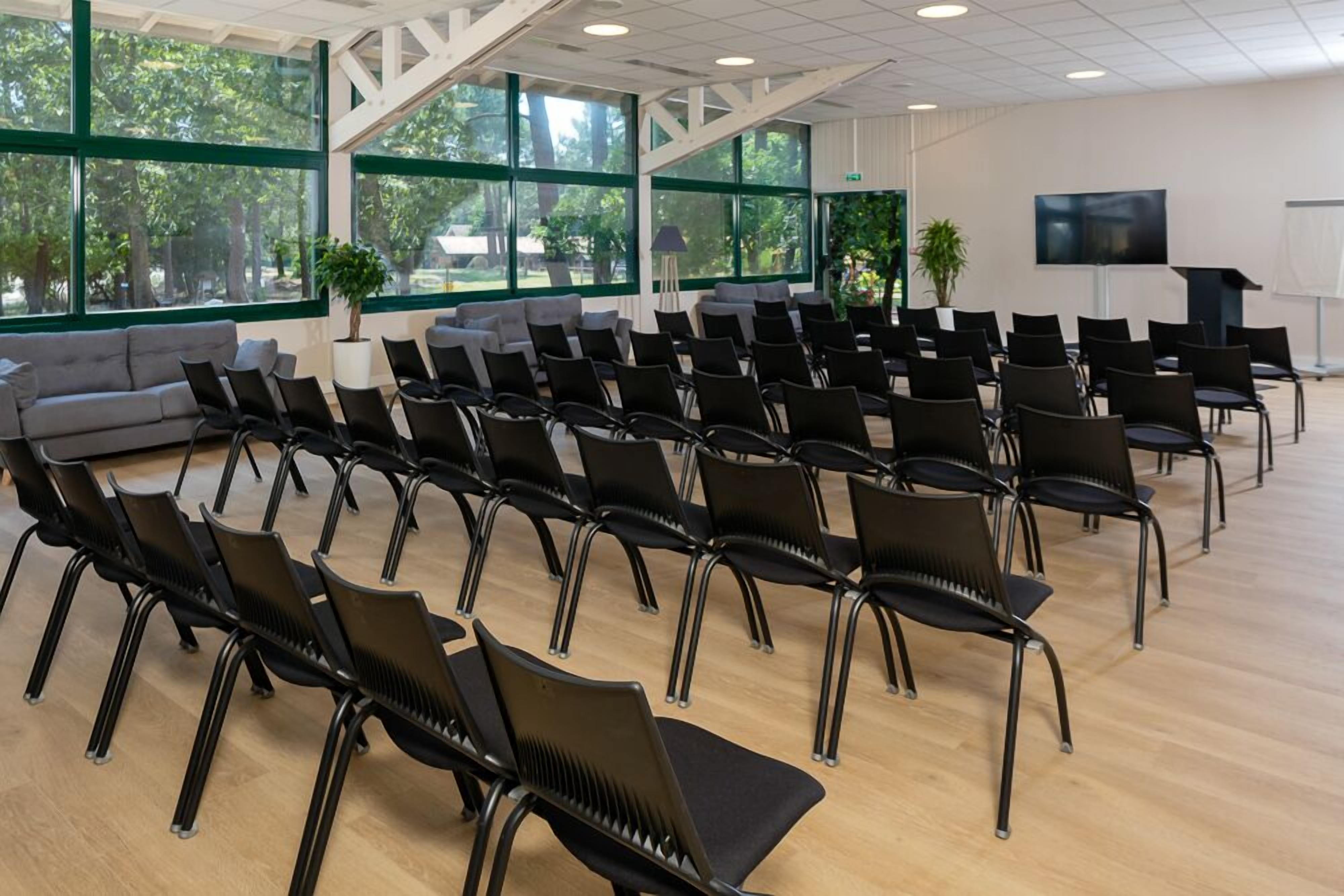 Une salle de conférence avec des rangées de chaises noires, de grandes fenêtres aux montures vertes, un canapé et un écran de télévision.