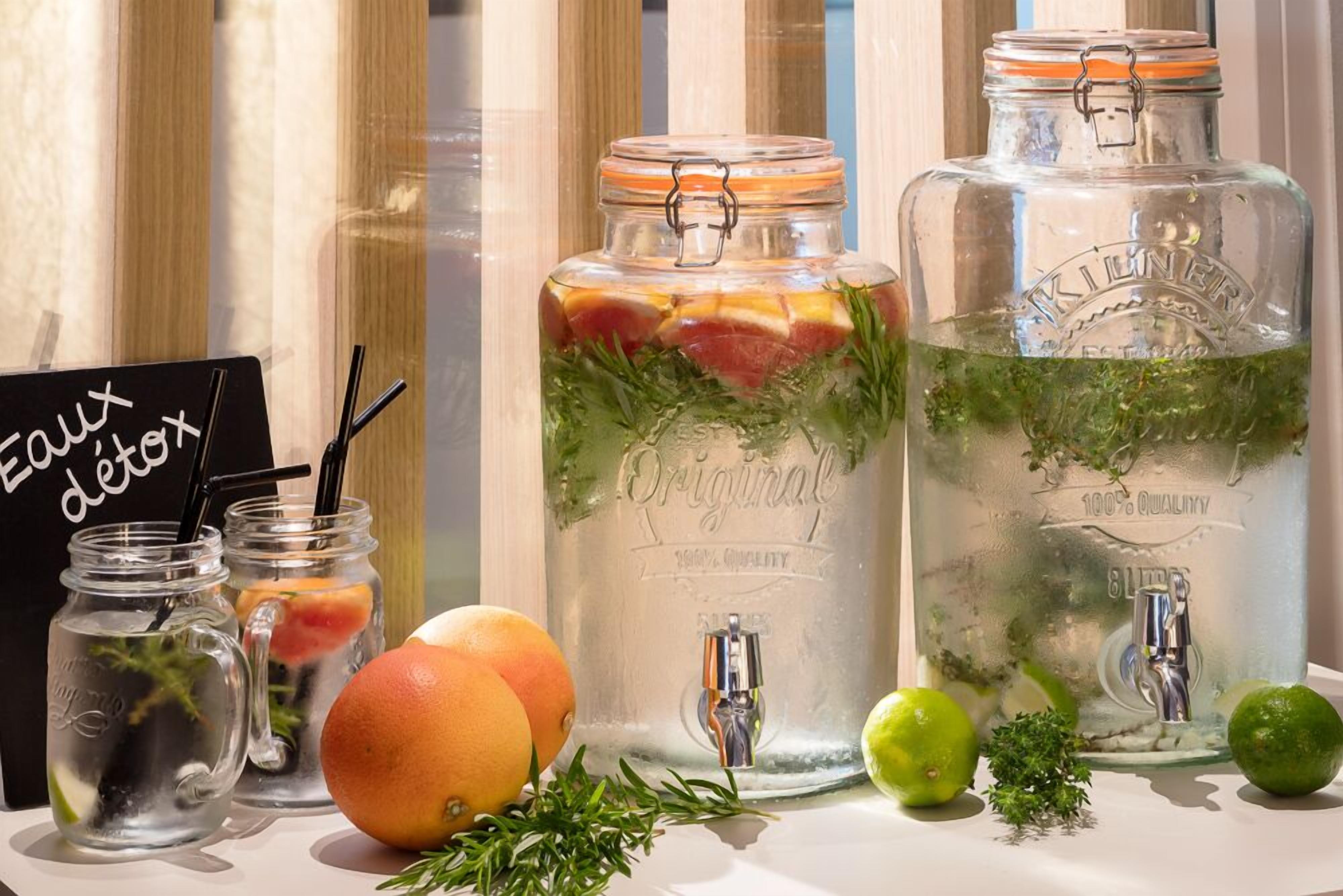 Deux grands bocaux en verre, dotés de robinets métalliques, sont remplis d’eau et de divers fruits et herbes. À côté, on trouve des bocaux plus petits, équipés de pailles, et un panneau indiquant « Eaux détox ».