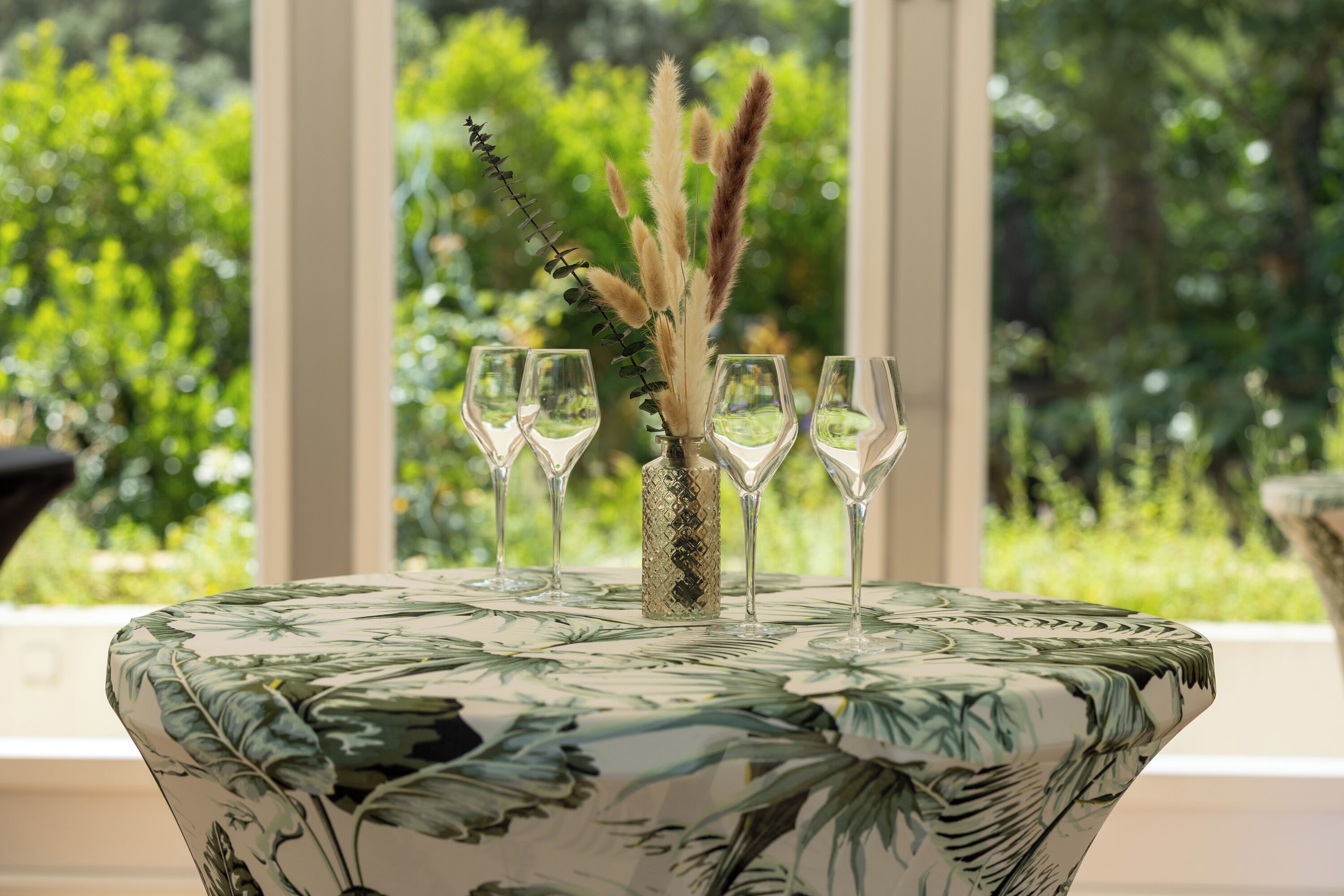 A table with a floral tablecloth, a vase with pampas grass, and four empty wine glasses.