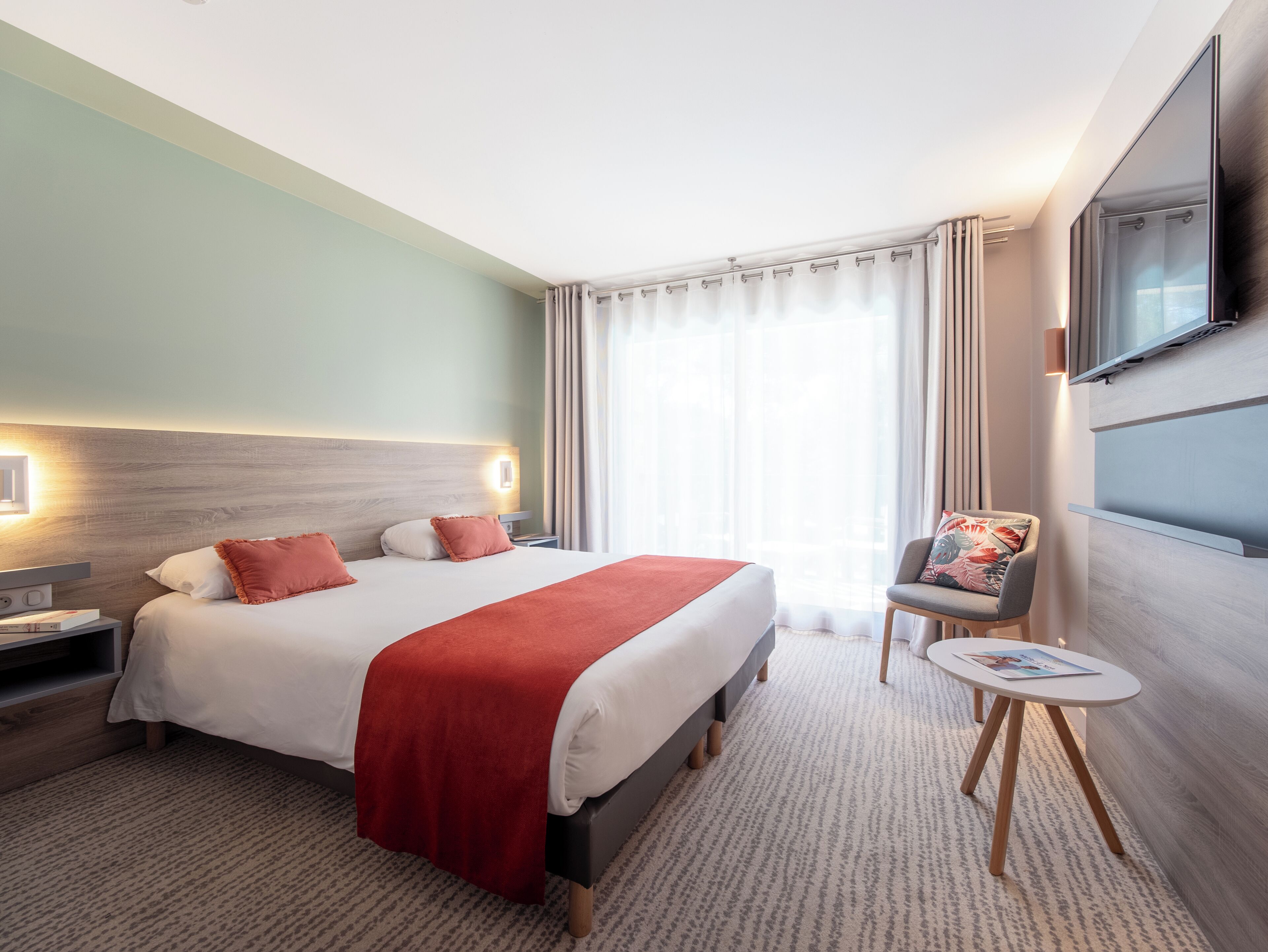 A hotel room with a bed, bedside table, chair, TV, and a window with curtains.
