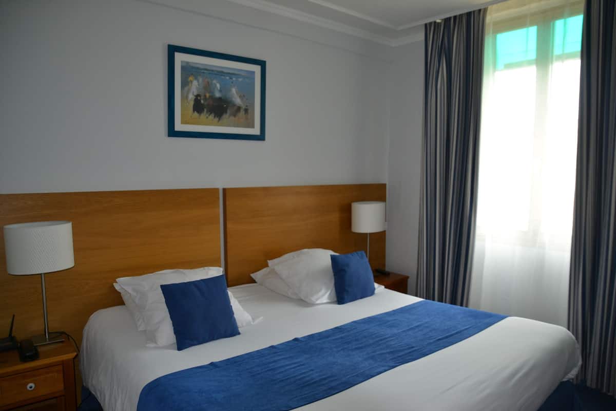 Habitación de hotel con una cama grande, dos lámparas de noche, un cuadro en la pared y una ventana con cortinas.