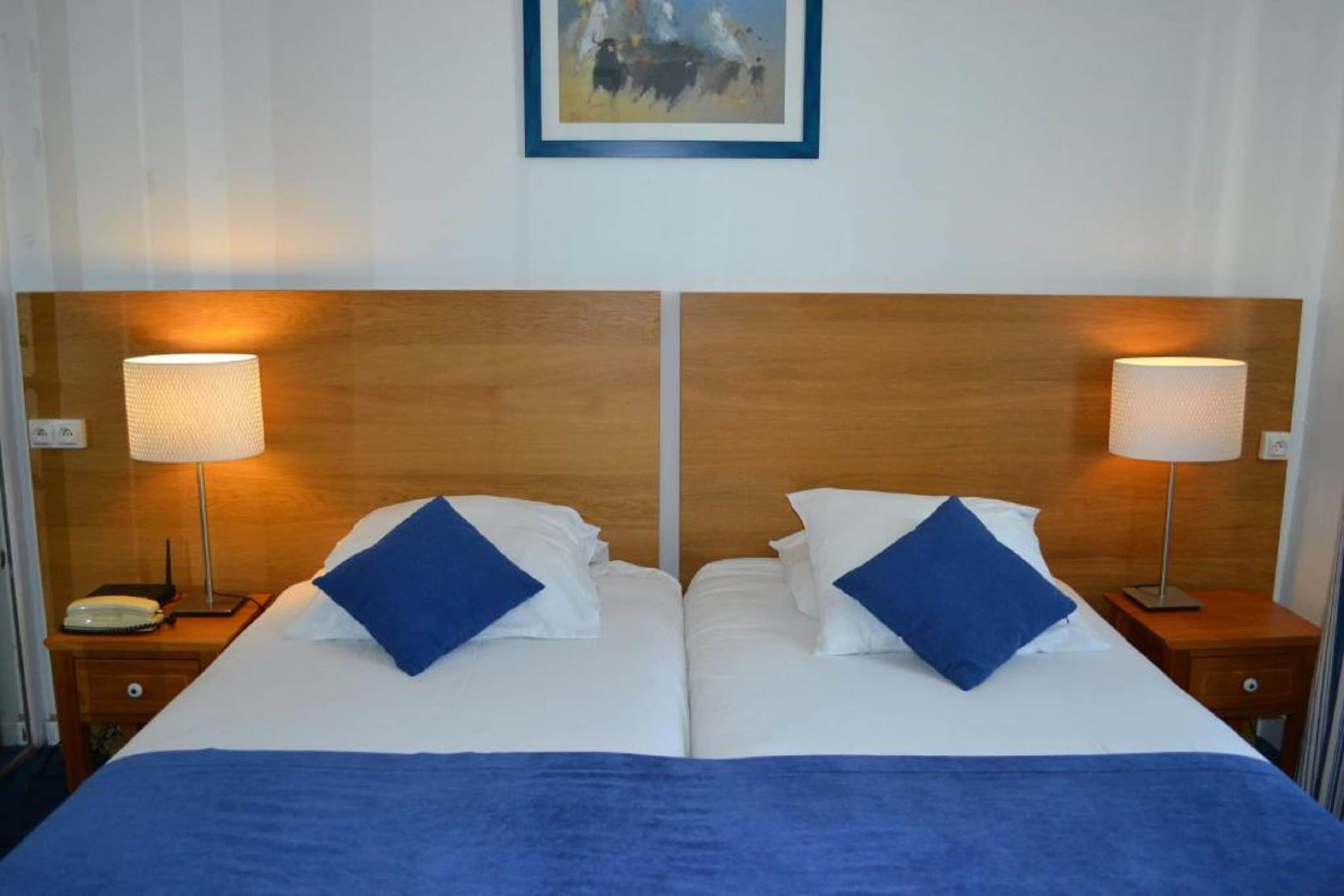 A double bed with white linens and blue pillows, flanked by wooden nightstands with lamps, in a room with a framed painting above.