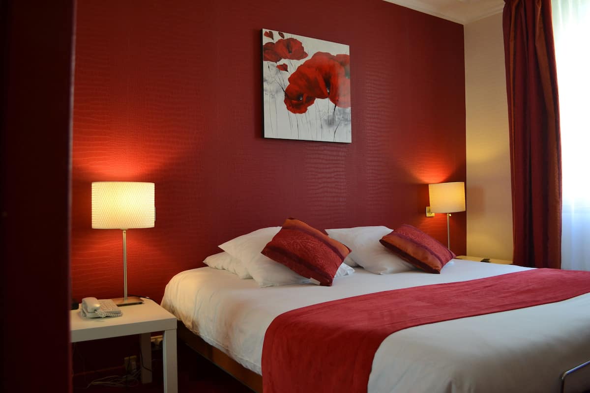 Habitación de hotel con pared roja, cama con ropa de cama blanca y almohadas rojas, dos lámparas de noche y un cuadro sobre la cama.