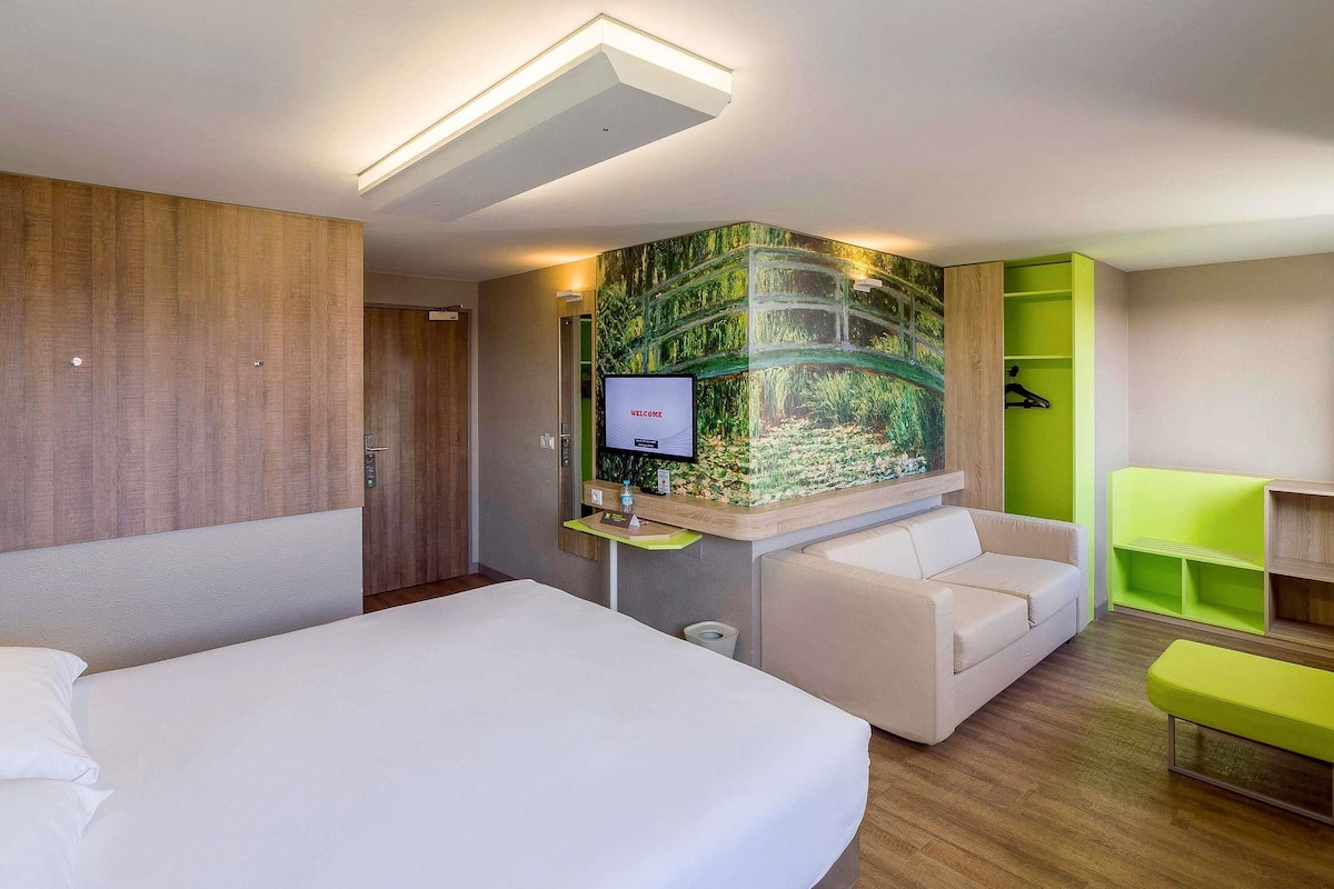 A hotel room with a bed, a sofa, a desk with a computer, a green storage unit, and a wall-mounted TV displaying 'WELCOME'.