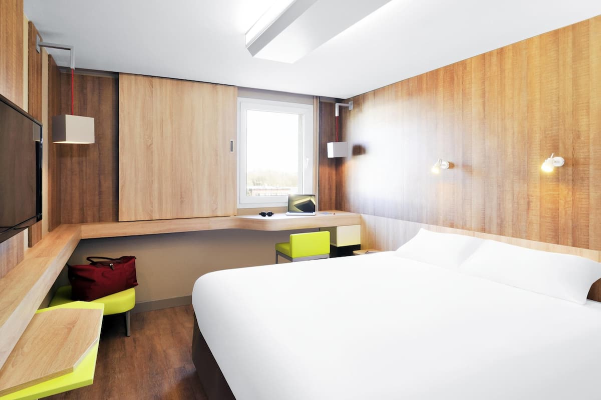 A modern hotel room with a bed, a desk, a chair, a TV, and a window.