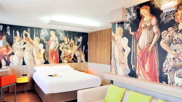 A hotel room with a bed, a desk, a white sofa, and a large mural on the wall. - ibis Styles Lille Marcq en Baroeul (Marcq-en-Baroeul)