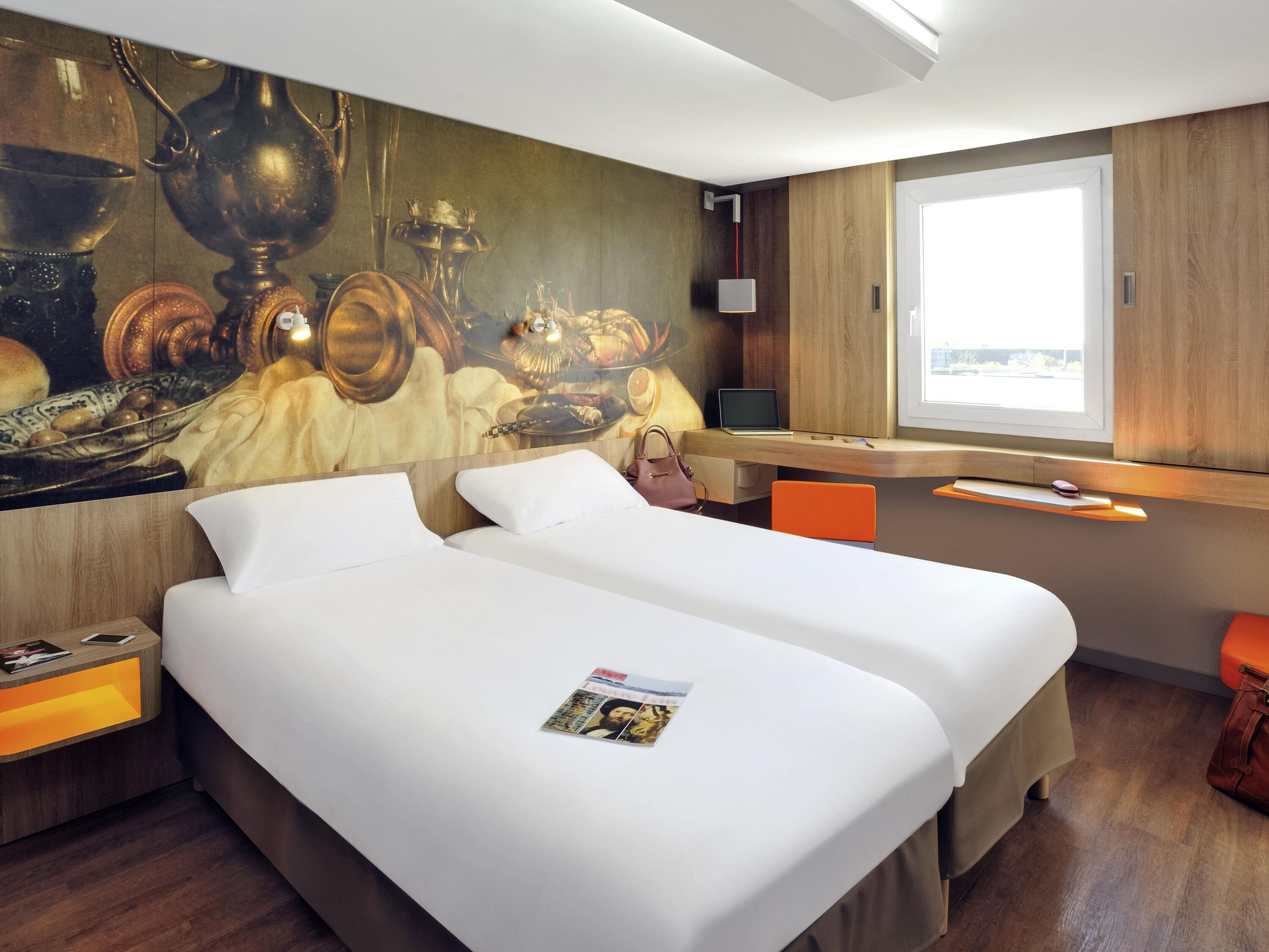 Une chambre d’hôtel avec deux lits, un bureau avec un ordinateur portable, une fenêtre donnant sur un paysage agréable et un mur orné d’un tableau.