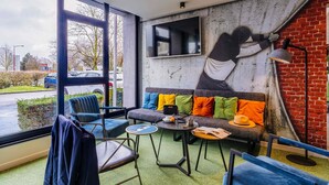 A modern lounge area with a large window, a mural, and a brick wall. - ibis Styles Lille Marcq en Baroeul (Marcq-en-Baroeul)