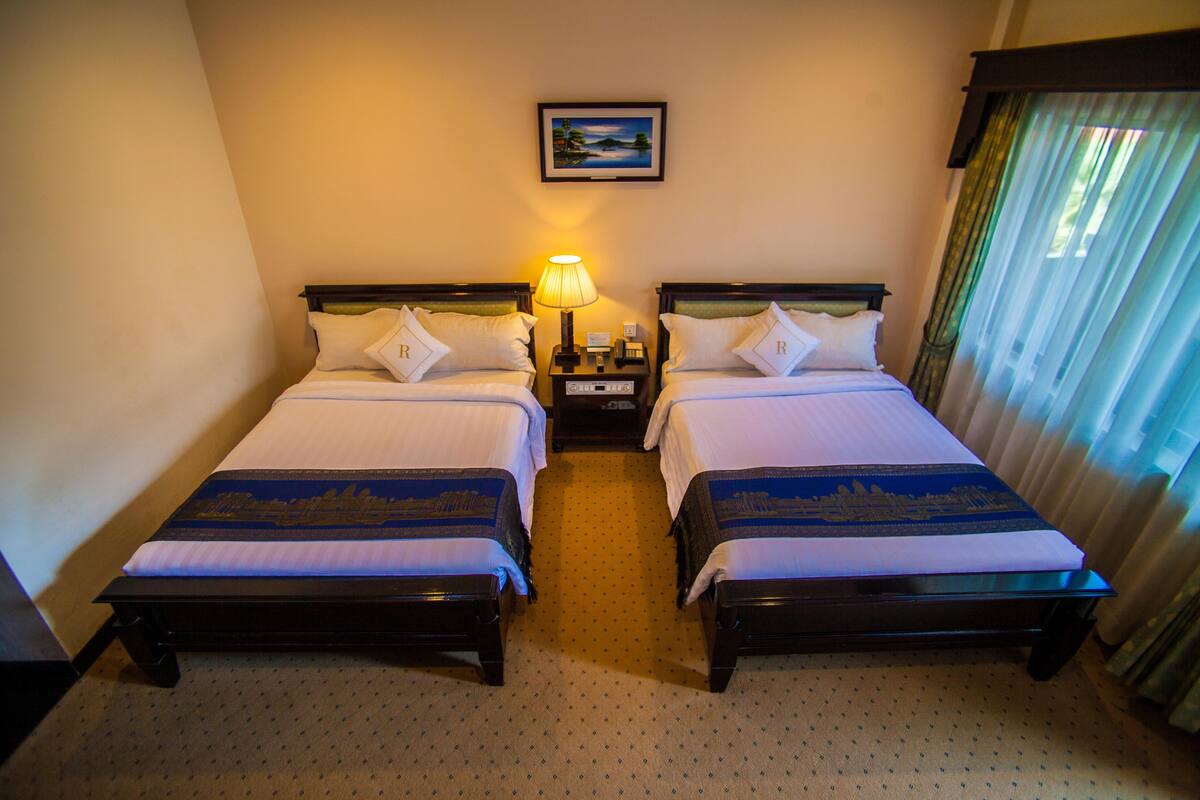 A hotel room with two single beds, a nightstand with a lamp, a painting on the wall, and a window with curtains.