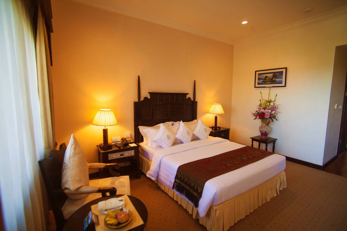 A hotel room with a large bed, two bedside tables with lamps, a chair, a small table with fruit, and a painting on the wall.