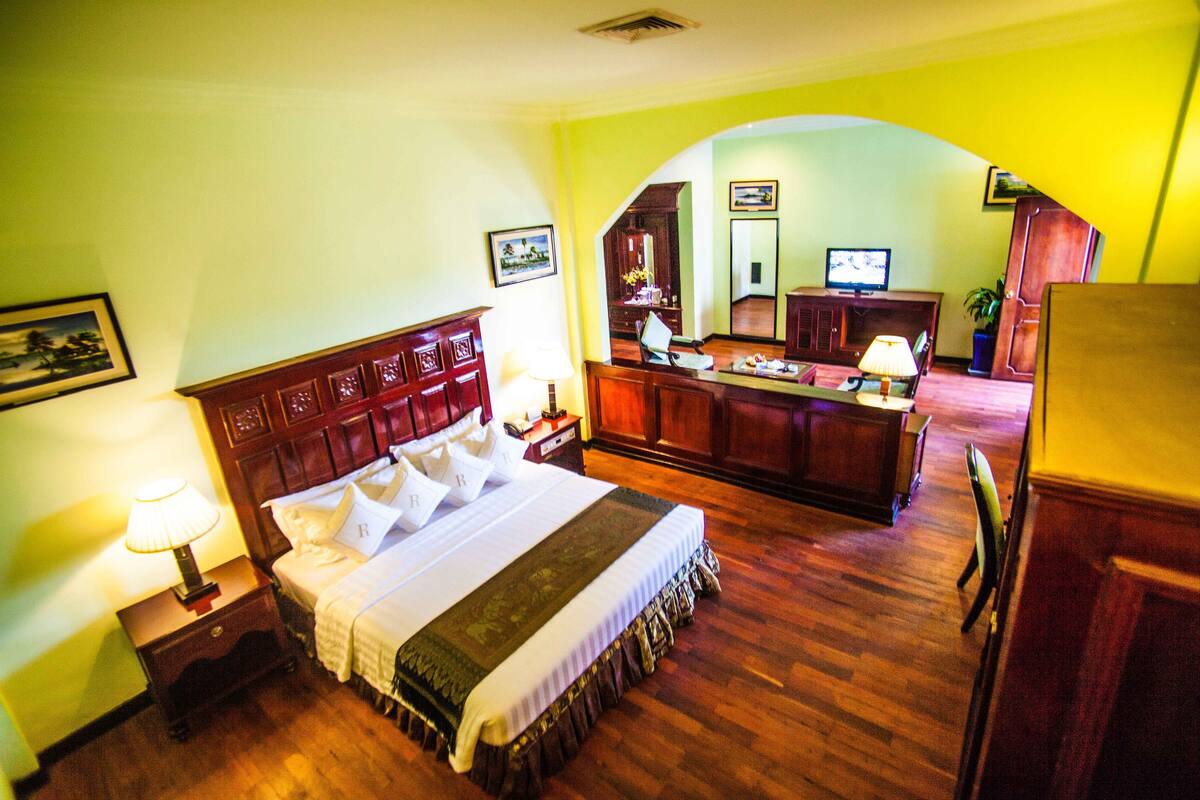 A hotel room with a large bed, wooden headboard, a desk with a chair, a television, and a small table with a lamp.