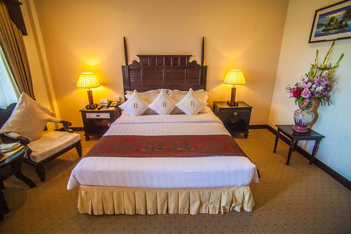 A hotel room with a large bed, two bedside tables with lamps, a chair, and a vase with flowers on a nightstand.