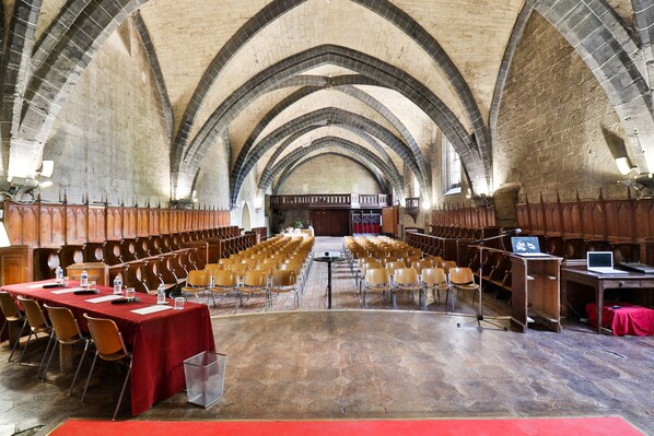Un gran salón con techos altos y arcos, filas de sillas de madera y mesas dispuestas para reuniones o conferencias.