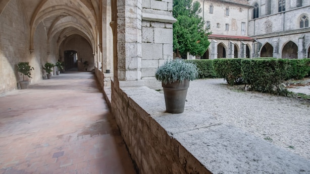 Un pasillo de piedra con arcos, plantas en macetas y un edificio con varias ventanas al fondo.