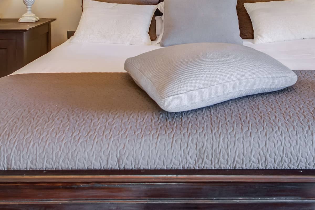 Una cama bien hecha con cabecera de madera, una lámpara blanca en una mesita de noche y una colcha blanca con textura.